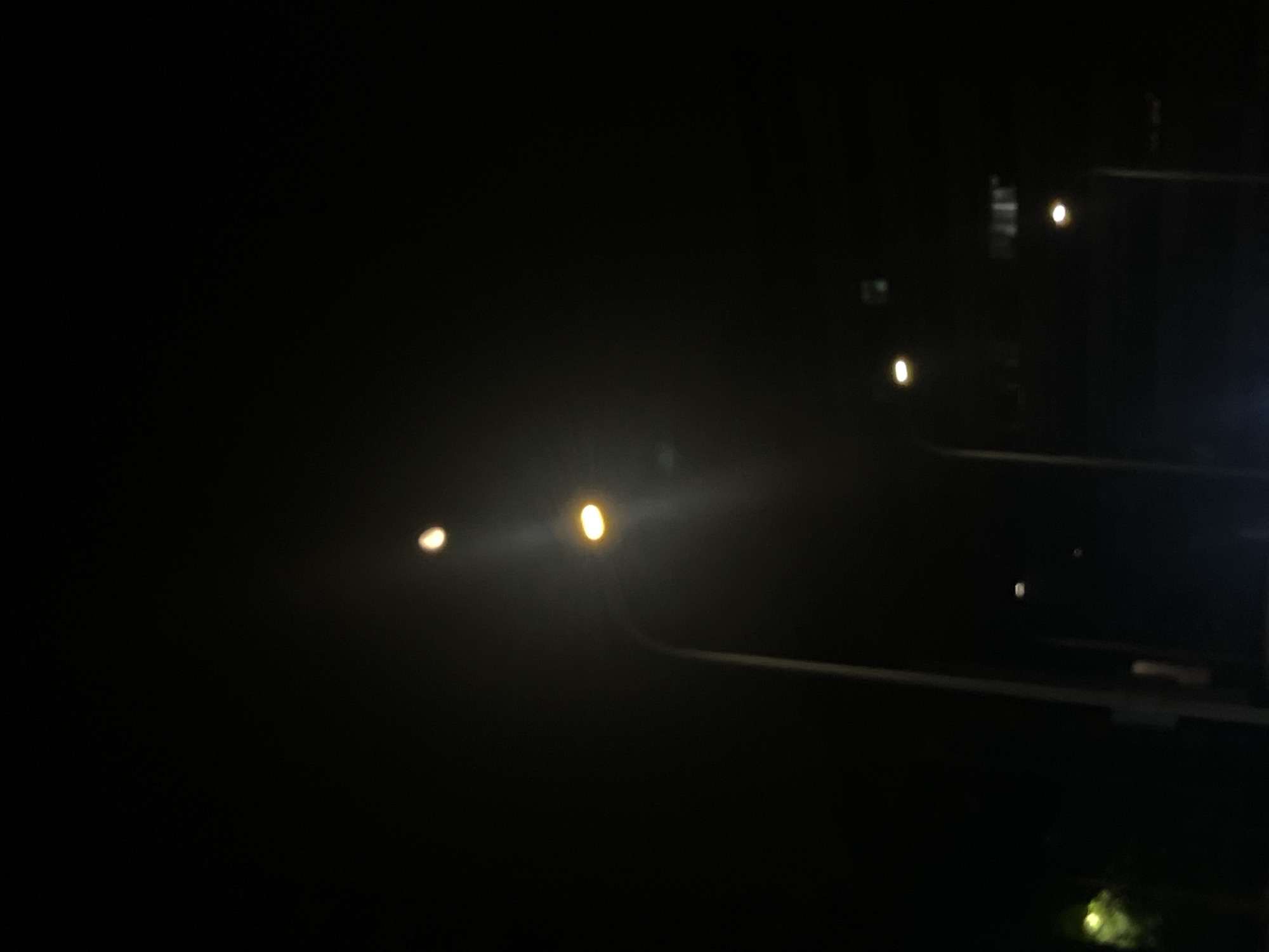 This image depicts a nighttime street scene within Yuzhong District, China. The environment is profoundly dark, with artificial light sources providing the primary illumination. A prominent street light, featuring a vertical pole, stands on the mid-left, emanating a bright yellow-orange light. A second, similarly illuminated street light is positioned further back and to the right. A distinct, smaller, yellowish light source is visible in the upper-center of the frame, separate from the primary street lights. Indistinct dark structures are faintly discernible in the far background on the right. A subtle green illumination is present in the lower-left quadrant. No individuals or active interactions are observed.