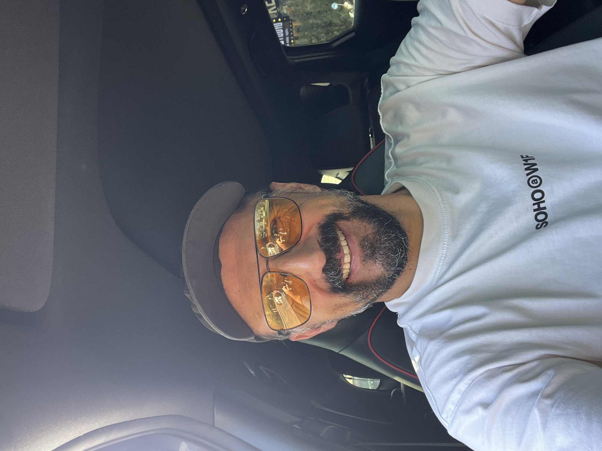 A man is inside a car in Baan Bo Phut, Thailand. The reflection in his amber-tinted glasses shows a street scene with palm trees. He's wearing a light-colored cap and a white shirt with the word "SOHO" printed on it. The interior of the car is black with red accents visible on the seats. A portion of the sunroof is visible.