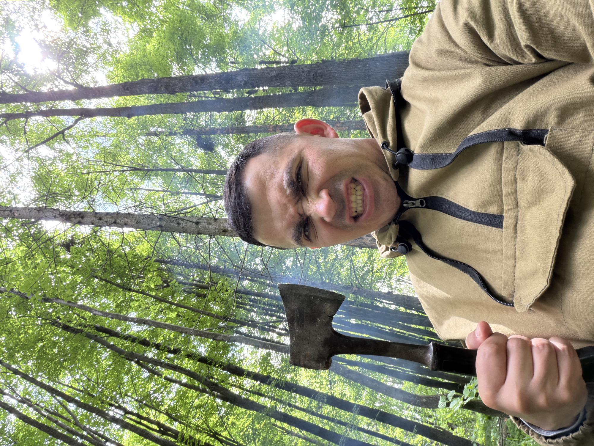 This is a low-angle, outdoor portrait taken in a forest environment in Kechur, Russia. The central figure is an adult male, likely in his 20s or 30s, with short dark hair and a clean-shaven face. He exhibits a strong grimace, with bared teeth and narrowed eyes, conveying an intense or aggressive expression. He wears a khaki-colored anorak-style jacket featuring a partial black zipper and a large front flap pocket. In his right hand, he firmly grips a rustic, weathered axe with a dark handle and a visibly worn metal head. The background consists of numerous tall, slender trees with abundant green foliage, suggesting a dense woodland. Sunlight filters through the tree canopy, creating bright areas. Wisps of light gray smoke are discernible amidst the trees in the background, indicative of a nearby heat source or smoldering material.