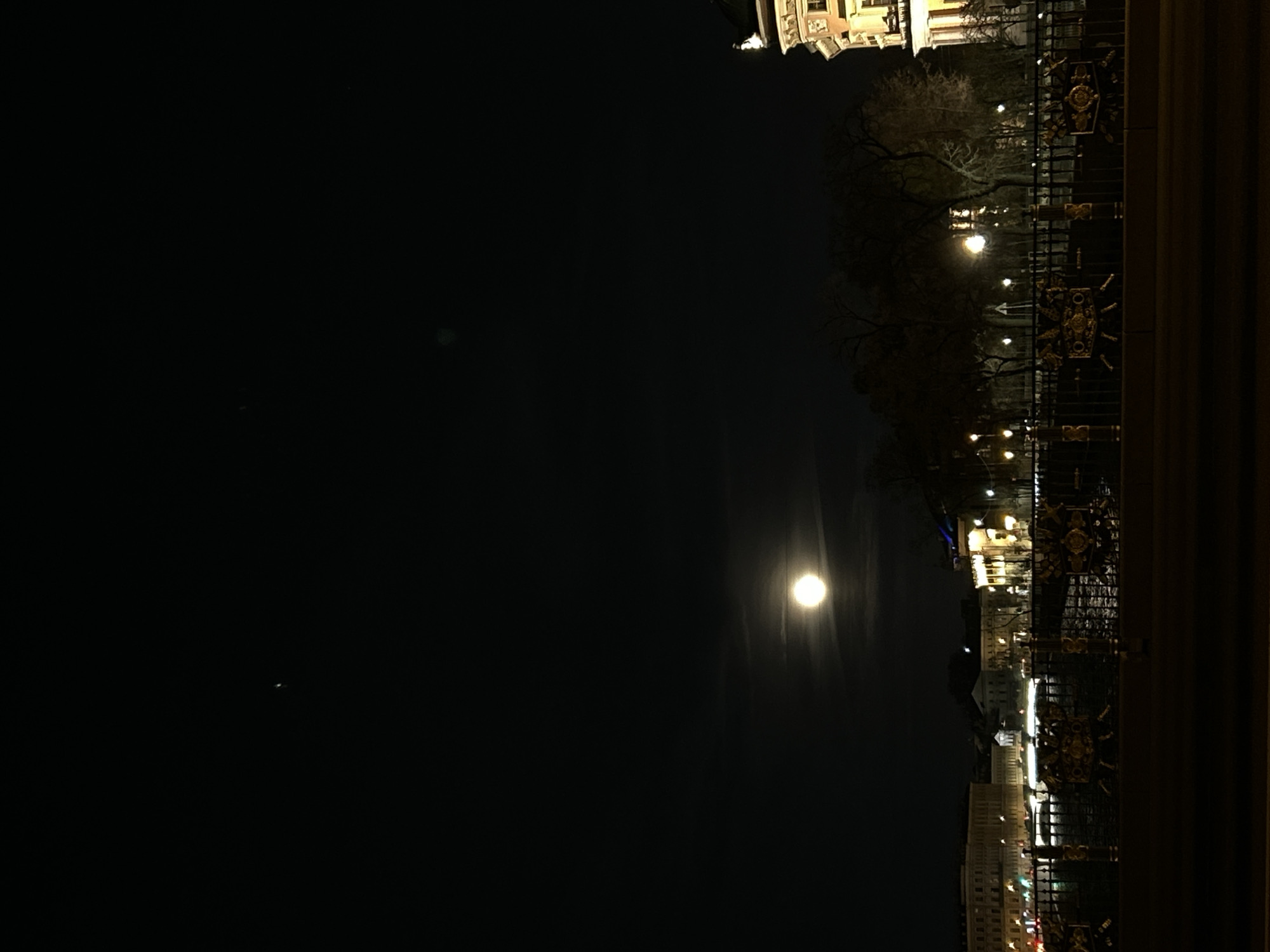 The image captures a serene nighttime scene in Saint Petersburg, Russia. The perspective is from a bridge or embankment, with a dark, solid structure and an ornate metal railing forming a foreground element at the top of the frame. This railing features dark bars with decorative golden details.
Below the foreground structure, a body of water, likely a river or canal, stretches across the midground, reflecting the numerous city lights. Along the opposite bank, buildings with classical architectural styles are illuminated, showcasing intricate details. Streetlights cast warm glows onto the scene, further defining the cityscape and silhouetting bare trees against the illuminated areas.
The dark, clear night sky dominates a large portion of the image. A very bright, seemingly full moon is prominently featured towards the middle-right of the frame, emitting a strong, radiant glow with visible light streaks emanating from it (likely lens flare). A few faint, blurry spots are also scattered in the vast darkness of the sky.
The overall impression is a quiet urban landscape at night, characterized by the clear sky, the bright moon, and the illuminated historic architecture of Saint Petersburg reflecting on the water. No specific activity or visible text is present.