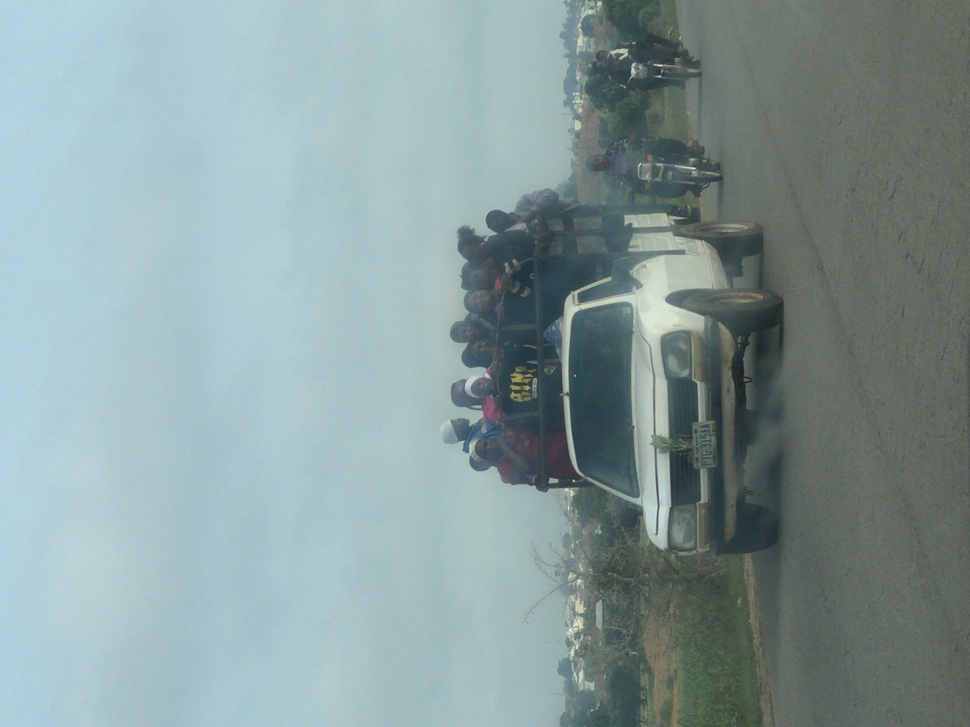 A medium shot captures a white pickup truck, laden with approximately 20 people, driving on a paved road. The truck's bed is filled with individuals, some standing and others sitting. In the foreground, a plant is visible growing from the front of the truck. Behind the truck, two motorcycles are also on the road, each with a rider. The background depicts a hazy, distant cityscape with buildings and trees. The sky is overcast. The license plate on the truck reads "PTG-315AA". The scene appears to be in Barakin, Nigeria, judging by the context provided.