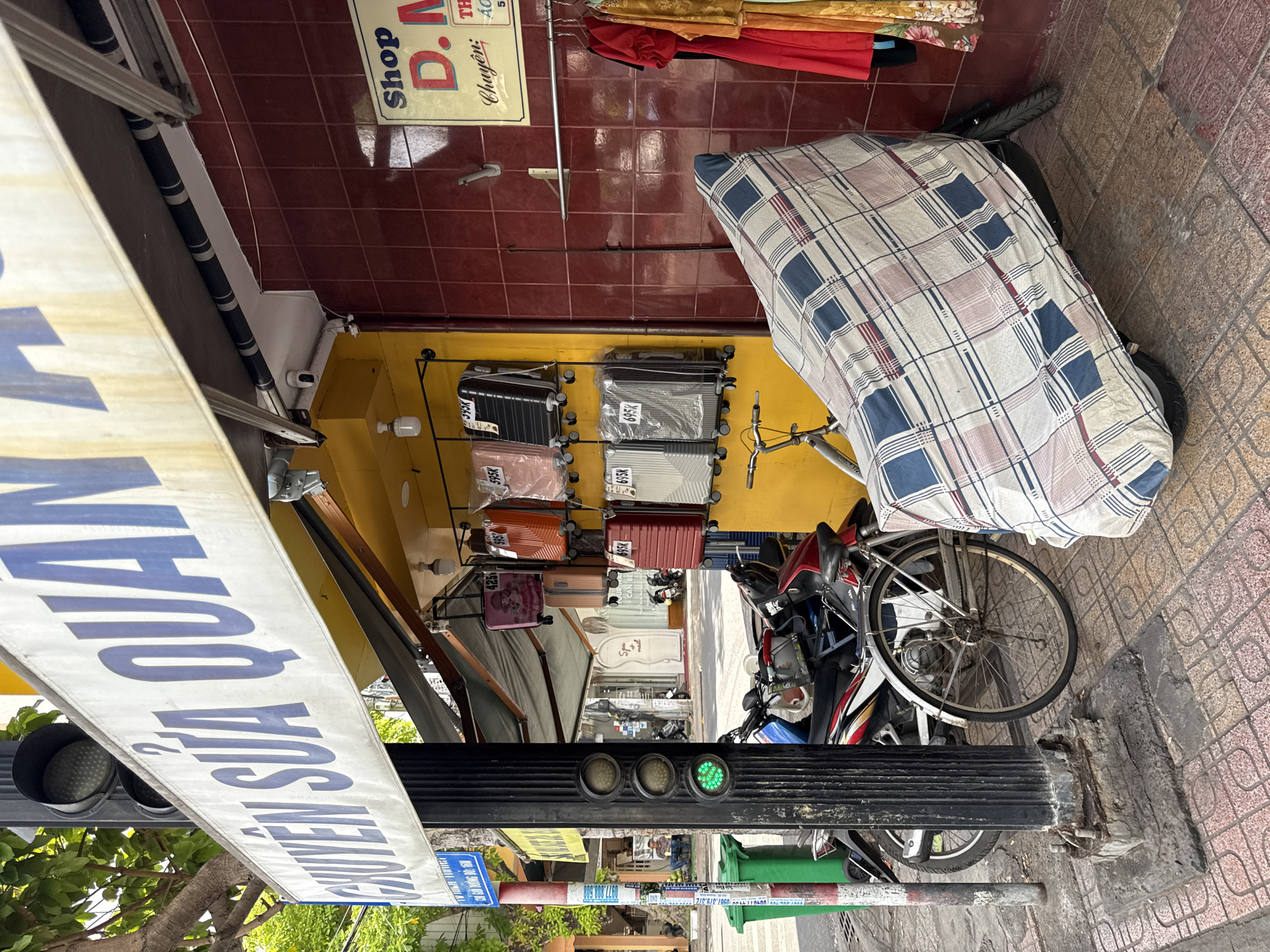The image depicts a street scene in Nha Trang, Vietnam. On the left, a traffic light with a green light illuminated is attached to a black ribbed pole. Next to the pole are two green trash bins. A banner with blue lettering hangs above the scene, reading "CHUYEN SỬA QUẠT..." (Specializing in fan repair...).
The main subject is a shopfront. A yellow wall forms the backdrop of the shop, displaying several suitcases of various colors and sizes arranged on shelves. Above the suitcases, a sign reads "Shop D.M.". To the right of the shopfront, a red tiled wall has clothes hanging on a rack, including a red dress and a yellow patterned garment.
In the foreground, a motorcycle is parked, covered with a blue and white plaid sheet. A bicycle is parked next to the motorcycle. The ground is paved with decorative tiles. In the background, a person is seated on a bench. The overall environment suggests a busy urban street with commercial activity.