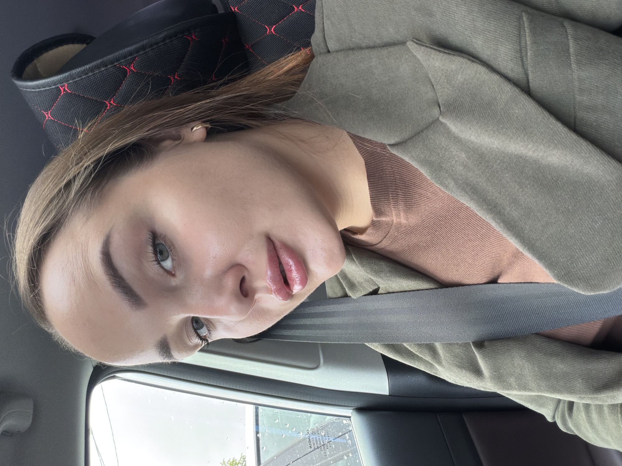 The image shows a young woman with light brown hair and green eyes, wearing a brown top and a green jacket. She is sitting in a car, with a seatbelt across her chest. The window on her left shows a blurry view of trees and possibly buildings, with rain streaks on the glass. The interior of the car includes a black car seat with red stitching. The woman is looking at the camera and appears to be taking a selfie. The scene is set in an urban environment, possibly on a rainy day in the city of Chaikovsky, Russia.
