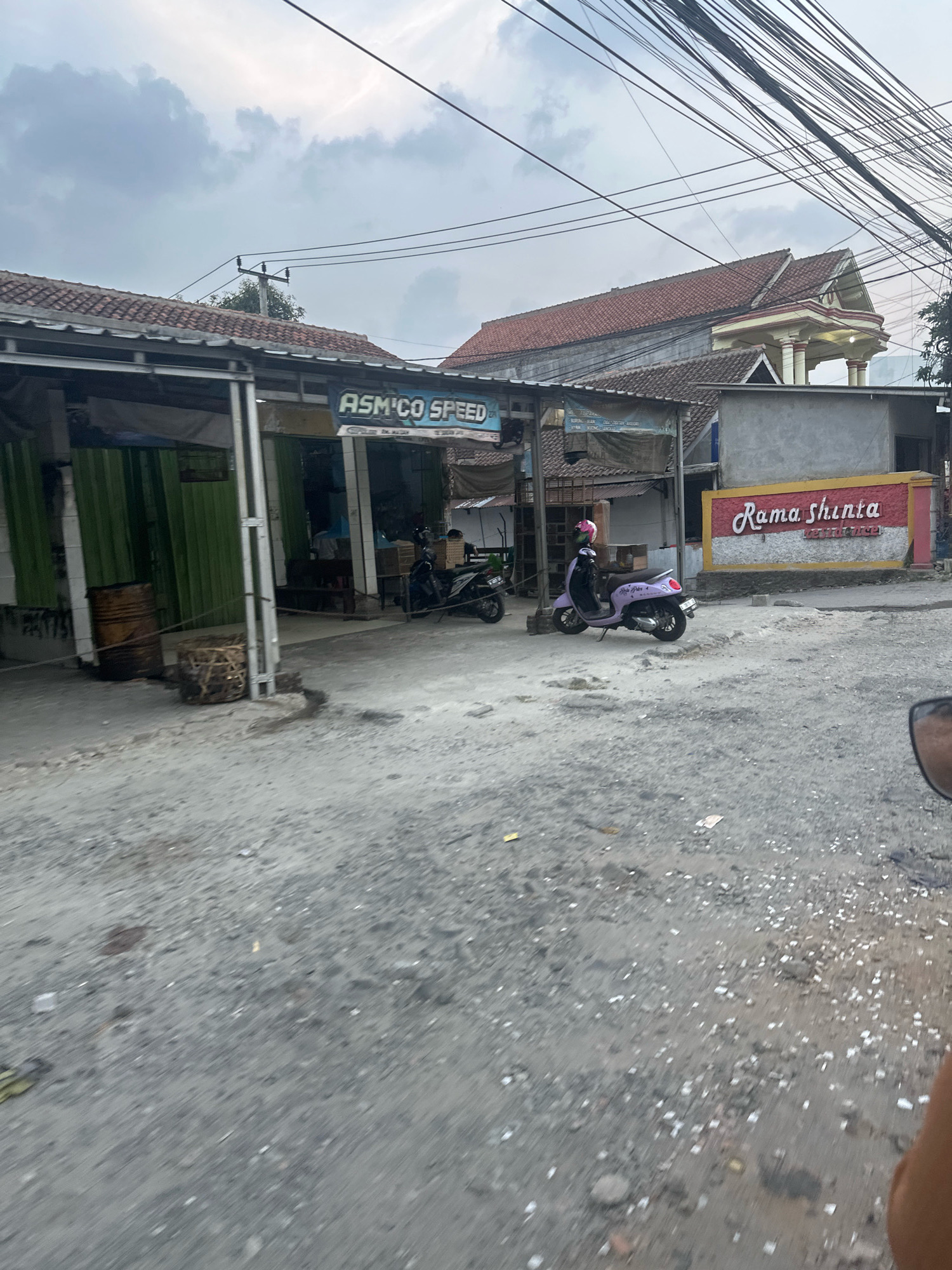 The image shows a street scene in Saruni, Indonesia. On the left, a shop with a green corrugated metal facade is visible, with a barrel and a woven basket outside. A sign above the shop reads "ASM CO SPEED". To the right, a purple scooter with a pink helmet is parked. Further to the right, a wall with a sign that says "Rama Shinta" in red and white lettering is present. In the background, a building with a red tiled roof and columns is seen. The street is unpaved and appears to be under a cloudy sky. Electrical wires are strung overhead. The image is taken from a moving vehicle, as indicated by the slight blur and the presence of a side mirror in the lower right corner.