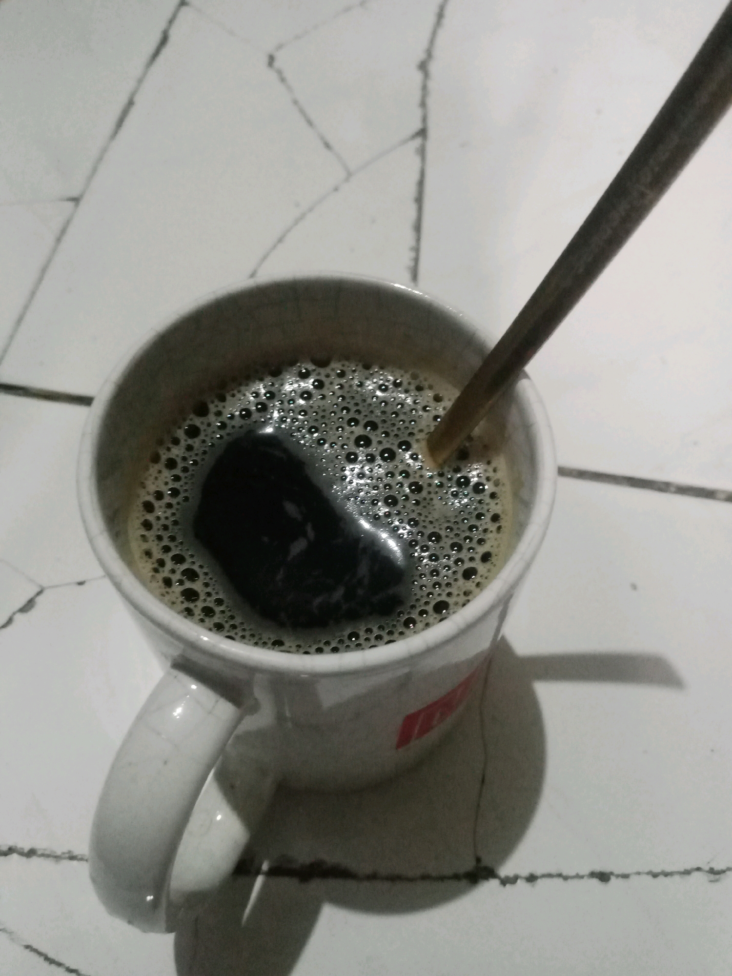 A white mug of coffee sits on a tiled surface with visible grout lines. A spoon is partially submerged in the coffee, which has a foamy surface and a dark, opaque appearance. The mug has a handle and a red logo partially visible on its side. Shadows are cast from the mug and spoon onto the tiled surface, indicating a light source from the top right. The tiles are light-colored with dark grout lines forming a geometric pattern. The overall scene is static and shows a simple beverage presentation. The location is in Balaraja, Indonesia.