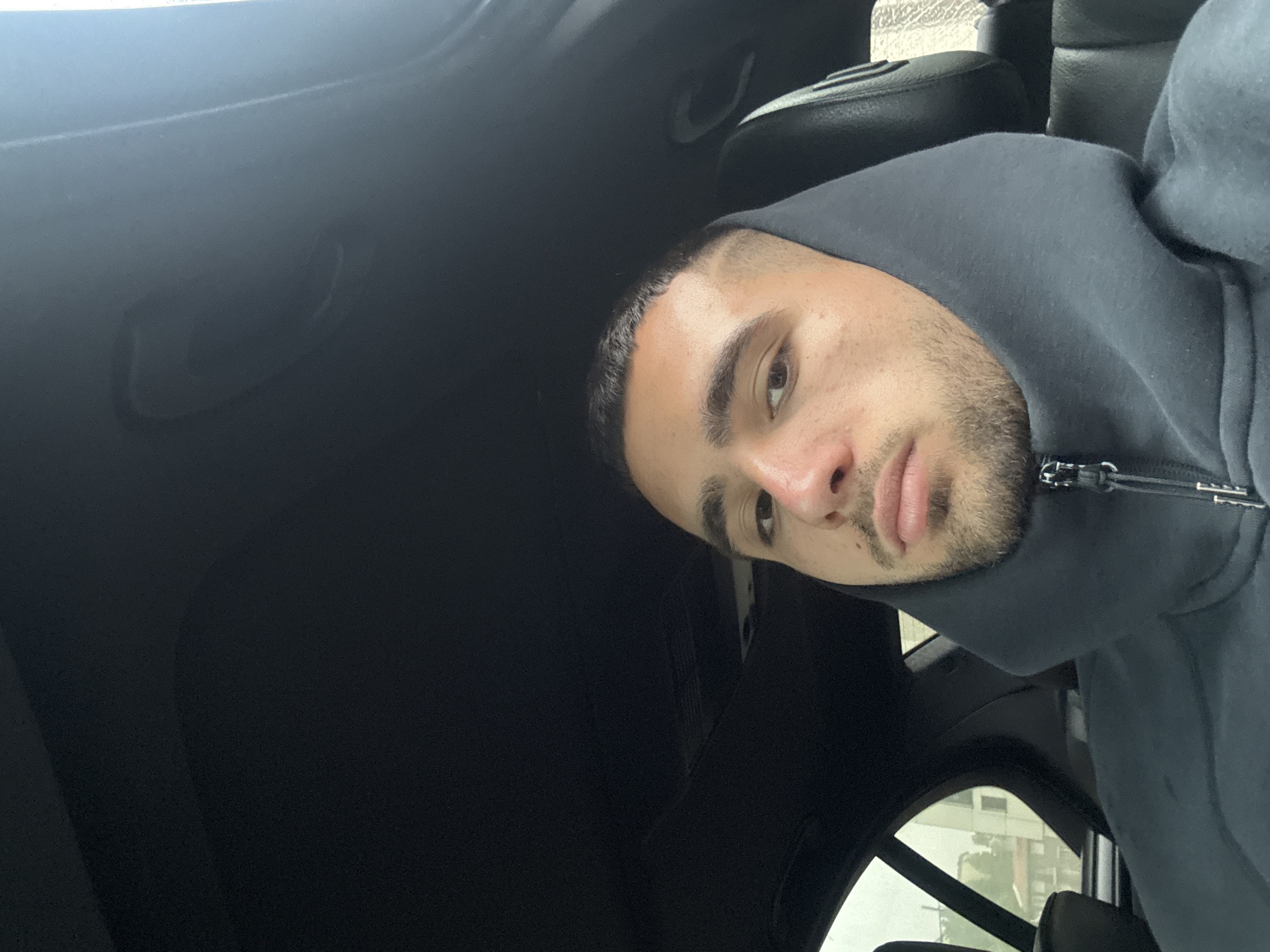 A man with short dark hair and a beard is sitting in a car, wearing a gray hoodie. He is looking at the camera. The car's interior is mostly black. Through the window, a building is visible. It is not possible to determine if the location is the city of Gabrovo, Bulgaria, based solely on this image.