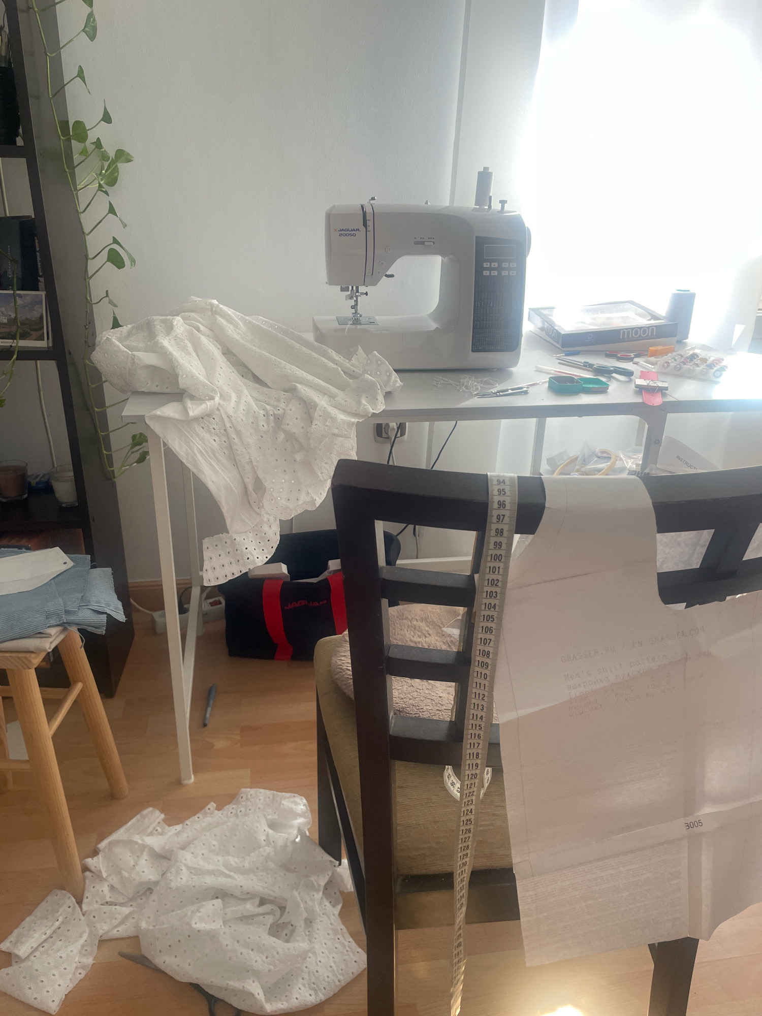 The image depicts a brightly lit room, likely a sewing room, with a strong light source coming from a large window on the right, causing some overexposure. In the center of the scene, a white sewing machine is positioned on a table. To the left of the sewing machine, a pile of white eyelet fabric is draped over a white folding table. Below this table, on the floor, are more pieces of the same white fabric and a pair of scissors. A wooden stool with stacked blue fabric is visible to the far left. A black bag with red trim, labeled "JAGUAR," is on the floor behind the folding table.
A wooden chair with a dark frame occupies the foreground, with a white piece of paper draped over its back. A measuring tape hangs down from the chair's back, showing numerical markings from 94 to 140. This paper appears to be a sewing pattern, with text visible, including "GRASSER.RU" and "Men's shirt pattern."
On the table, behind the sewing machine, are various sewing supplies, including spools of thread, scissors, pens, and a box labeled "moon." A houseplant with long vines is growing from a shelf unit on the far left of the image.
The floor is made of wooden planks. The overall impression is a workspace where sewing projects are in progress. There are no individuals present in the image. The location is Atarfe, Spain.