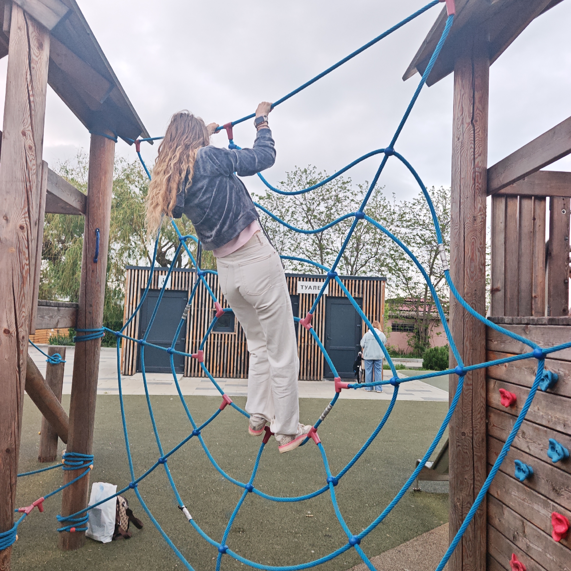 A girl with long, curly hair, wearing a blue jacket, pink shirt, and light-colored pants, is climbing on a blue rope net at a playground. She is facing away from the camera, holding onto the ropes with both hands and standing on the net. The playground has wooden structures, including a climbing wall with blue and red grips. In the background, there is a modern building with vertical wooden slats and the word "туалет" (toilet) written on it, suggesting it is a restroom facility. An adult, possibly supervising the girl, stands near the restroom. The ground of the playground is covered in green artificial turf. A bag and a plastic bag sit on the ground at the base of one of the wooden supports. The sky is overcast, indicating a cloudy day.