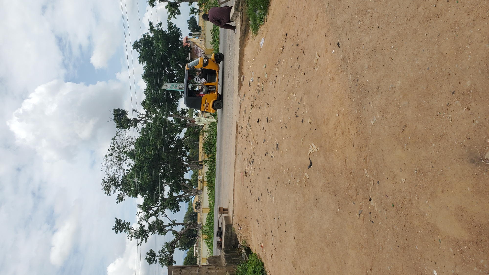 The image depicts a street scene in Jalingo, Nigeria, on a partly cloudy day. In the foreground, there is a dirt and gravel area with some scattered debris. A low concrete structure with a wooden plank is visible on the left. A road runs horizontally across the middle ground. On the road, a yellow auto-rickshaw, also known as a Keke Napep, is moving. It appears to be carrying passengers. To the right of the road, a person is seated on a concrete ledge. In the background, there are trees and some buildings, with a distant view of hills under a cloudy sky. A man in a light-colored garment stands on the side of the road near the auto-rickshaw.
