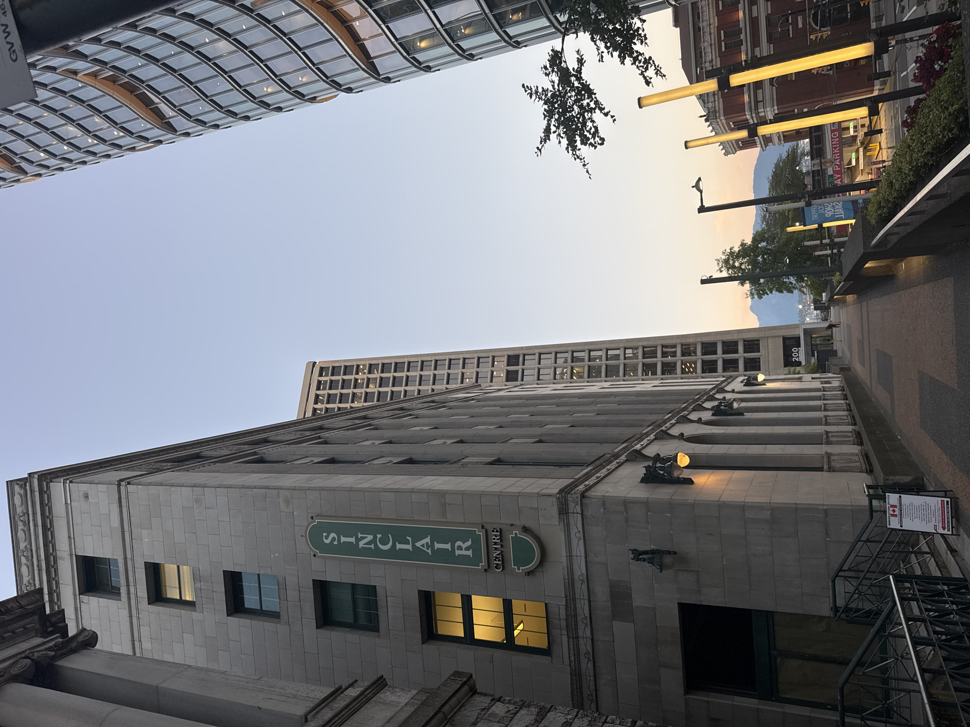 The image captures a street-level view of the Sinclair Centre, a federal government building located in Vancouver, Canada, at the intersection of Georgia Street and Granville Street. The Sinclair Centre, a heritage building, is visible on the left, characterized by its grey stone facade and distinctive green signage with "SINCLAIR CENTRE" written vertically. Adjacent to it, to the right, is a modern, curved glass skyscraper. In the background, another taller, rectangular building stands, and beyond that, mountains are visible under a pale sky, suggesting either early morning or late evening. The foreground shows a pedestrian walkway with a railing, leading towards the street. Several yellow vertical lights line the walkway, and a tree with dark green foliage is present on the right side of the frame. A sign for "DAY PARKING" is visible in the distance. The overall scene depicts an urban environment with a mix of historic and contemporary architecture.