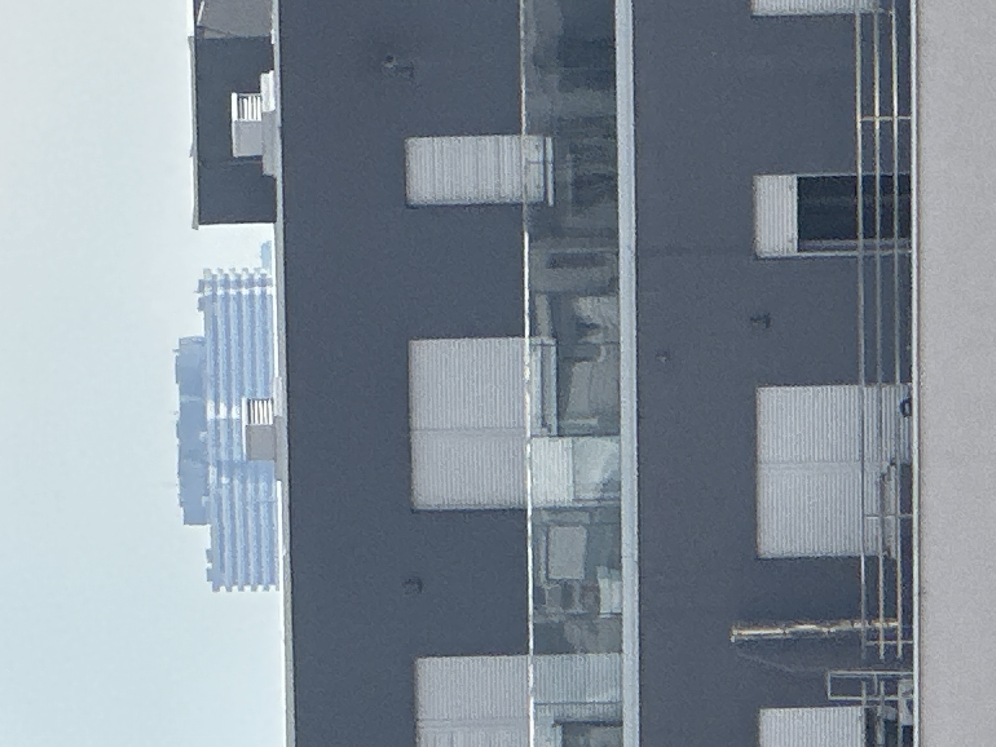This is a telephoto shot of a modern, multi-story building in Belgrade, Serbia, under a hazy, bright sky. The building's facade is a dark grey. On the first visible level from the bottom, there are white, slatted roller shutters covering what appear to be large windows or doors, and a metal railing runs along the front. Above this, a second level features similar white roller shutters, separated by a glass balcony railing. In the distance, a long, multi-story building with horizontal blue and white bands is visible, possibly another residential complex or office building. The overall impression is one of urban architecture with a focus on clean lines and functional design.