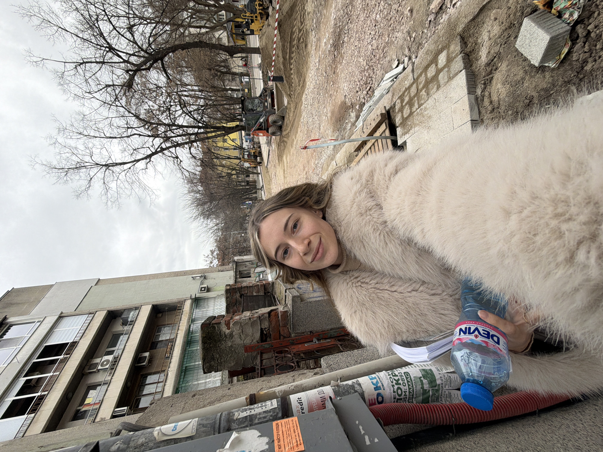 A woman wearing a large, light-colored fur coat is taking a selfie outdoors. In her left hand, she holds a bottle of Devin water. The background shows a cityscape with buildings, construction work including heavy machinery and a trench, and bare trees. The woman is positioned near construction materials, including pipes and items with Cyrillic writing. The OCR provides text that mentions "My master in Plovdiv" and a GSM number, suggesting the location is Plovdiv, Bulgaria. The overall impression is a casual selfie taken in an urban environment, possibly near an area undergoing construction.