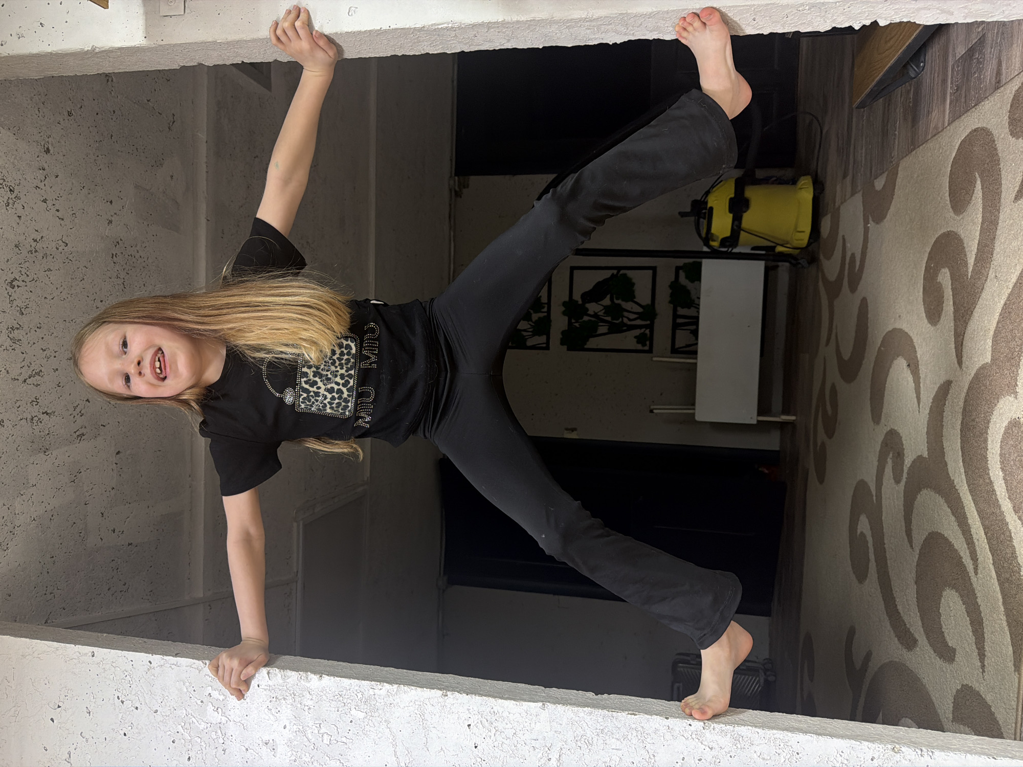 A young blonde girl, dressed in a black t-shirt featuring a "WILD CHILD" leopard print design and black leggings, is playfully suspended upside down within a rectangular opening of a building. Her hands and bare feet grip the textured, light-colored concrete or plaster surfaces of the opening, forming a stretched "X" shape with her body. She smiles broadly towards the camera, conveying joy and playfulness.
The scene is indoors, within a building in Chaikovsky, Russia. Visible through the opening behind the girl are elements of a domestic setting: dark wooden flooring, a beige and brown patterned rug with a swirling design, and various household objects. These include a bright yellow portable cooler or container with straps, a white table or counter, and dark-framed artwork or decorations on a wall, possibly depicting abstract trees or plants. The lighting is ambient, typical of an indoor environment. The textured, concrete-like walls suggest a modern or possibly industrial-inspired interior design within a home or similar structure.