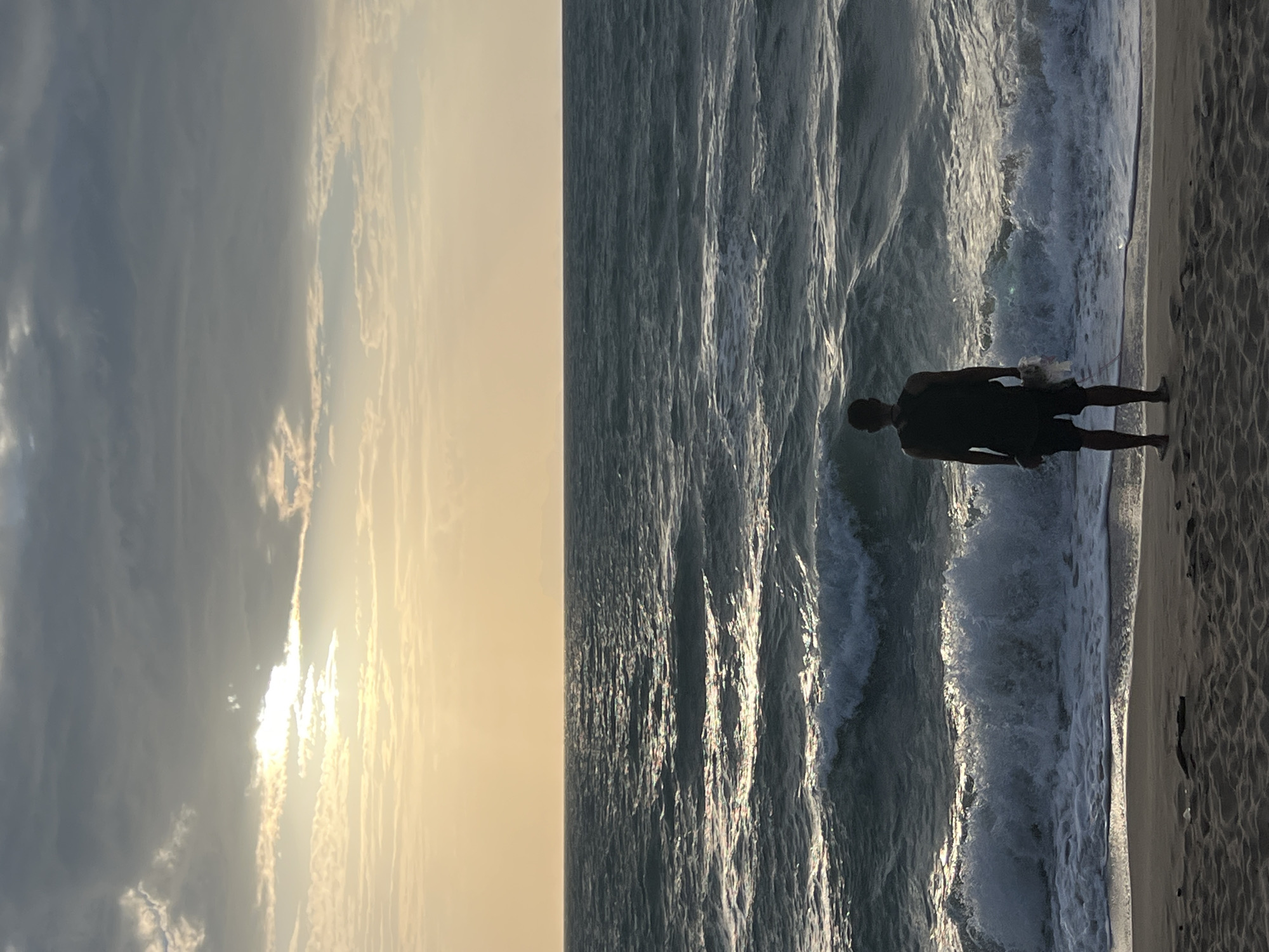 A person stands on a sandy beach facing a rough, choppy sea under a cloudy, overcast sky with the sun peeking through the clouds. The person is wearing a dark tank top, dark shorts, and a backpack. They are holding a white plastic bag in their right hand. The waves are breaking near the shore, creating white foam. The sand on the beach is dark. The horizon line is visible in the distance, with a faint outline of land. The scene appears to be at dusk or dawn, with the sky illuminated by the setting or rising sun. The location is the coast of Ko Samui District, Thailand.