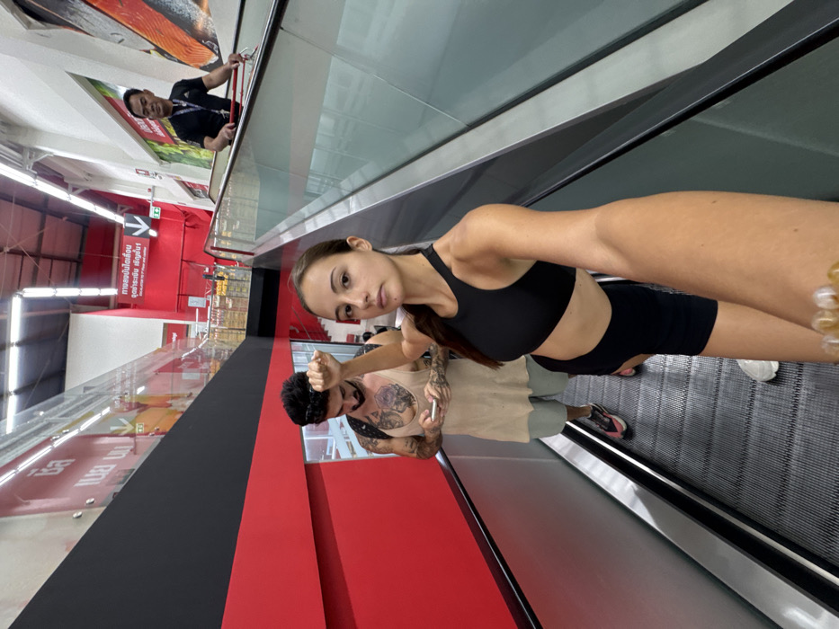 A low-angle selfie, taken on an ascending escalator inside a Big C Supercenter in Bangkok, Thailand, captures three individuals. In the immediate foreground, a young adult female with light skin and brown hair, wearing a black sports bra and black cycling shorts, extends her right arm to operate the camera, displaying a serious expression. Her left arm is raised, with the hand clenched into a fist positioned near her head. Behind her, on the same escalator, stands a young adult male with dark hair and a beard, adorned with visible tattoos on his chest and arms. He wears a beige sleeveless tunic over light shorts and athletic shoes, engrossed in a white smartphone held in both hands. Sunglasses are pushed onto his head. Further in the upper right background, an adult Asian male, likely a store employee in a black polo shirt with a lanyard, rides a parallel ascending escalator, holding the handrail. The store interior features prominent white walls with large red and black panels. Overhead, fluorescent lighting is visible. Thai script signage is present, including "บิ๊กซี" (Big C) on a red wall panel to the left, and a red sign on the right clearly indicating "Escalator to 2nd Floor and upstairs" (บันไดเลื่อนขึ้นชั้น 2 และชั้นบน). Merchandise shelving is visible on upper levels, and a large graphic of a salmon is displayed on a white wall section, indicating a fresh food area.