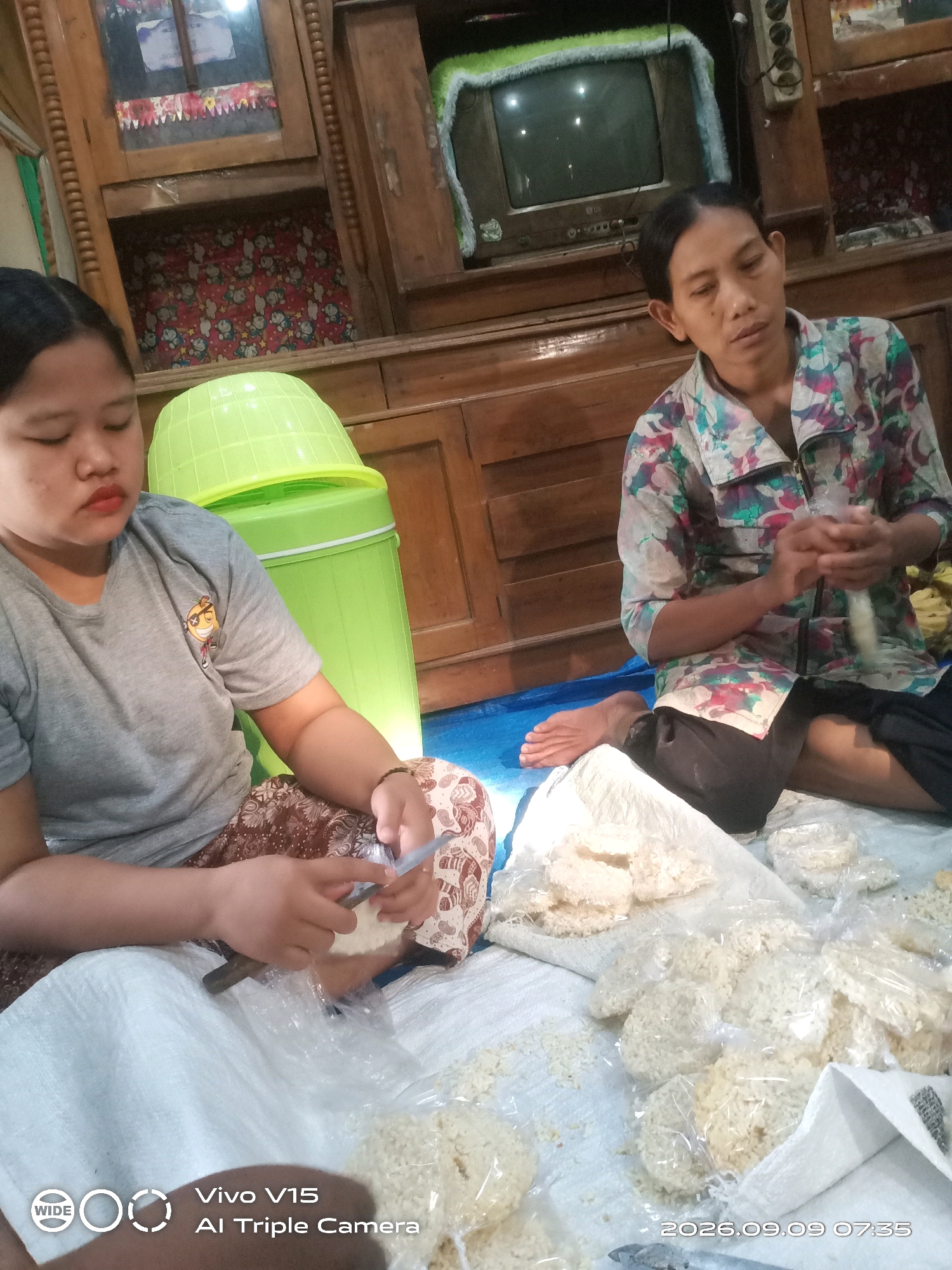 In a room with wooden cabinets and a television, two women are sitting on a blue tarp, preparing snacks. The woman on the left, wearing a grey t-shirt and a patterned skirt, is focused on her task, holding a small knife and what appears to be a piece of dough. To her right, a green trash can stands tall. The woman on the right, dressed in a floral shirt and dark pants, is also engaged in her work, her hands manipulating small white items. The floor is covered with white cloth and a variety of snacks, some individually wrapped in plastic. The environment appears to be a domestic setting, possibly in Plosorejo, Indonesia, indicated by the time stamp "2026.09.09 07:35" and the "Vivo V15 AI Triple Camera" watermark, often found on mobile devices used in the region.