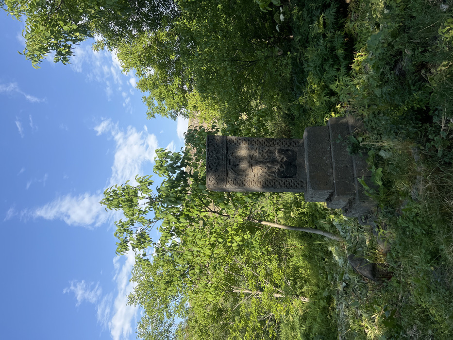 An ornate stone cross stands prominently among lush greenery, under a sky of soft blue and scattered clouds. The cross's intricate carvings contrast with the surrounding foliage. The structure is comprised of multiple stacked stone pieces. The verdant background enhances the cross's aged texture. Sunlight dapples through the canopy above, casting shadows on the historical monument. The scene evokes a sense of timeless beauty and serenity.