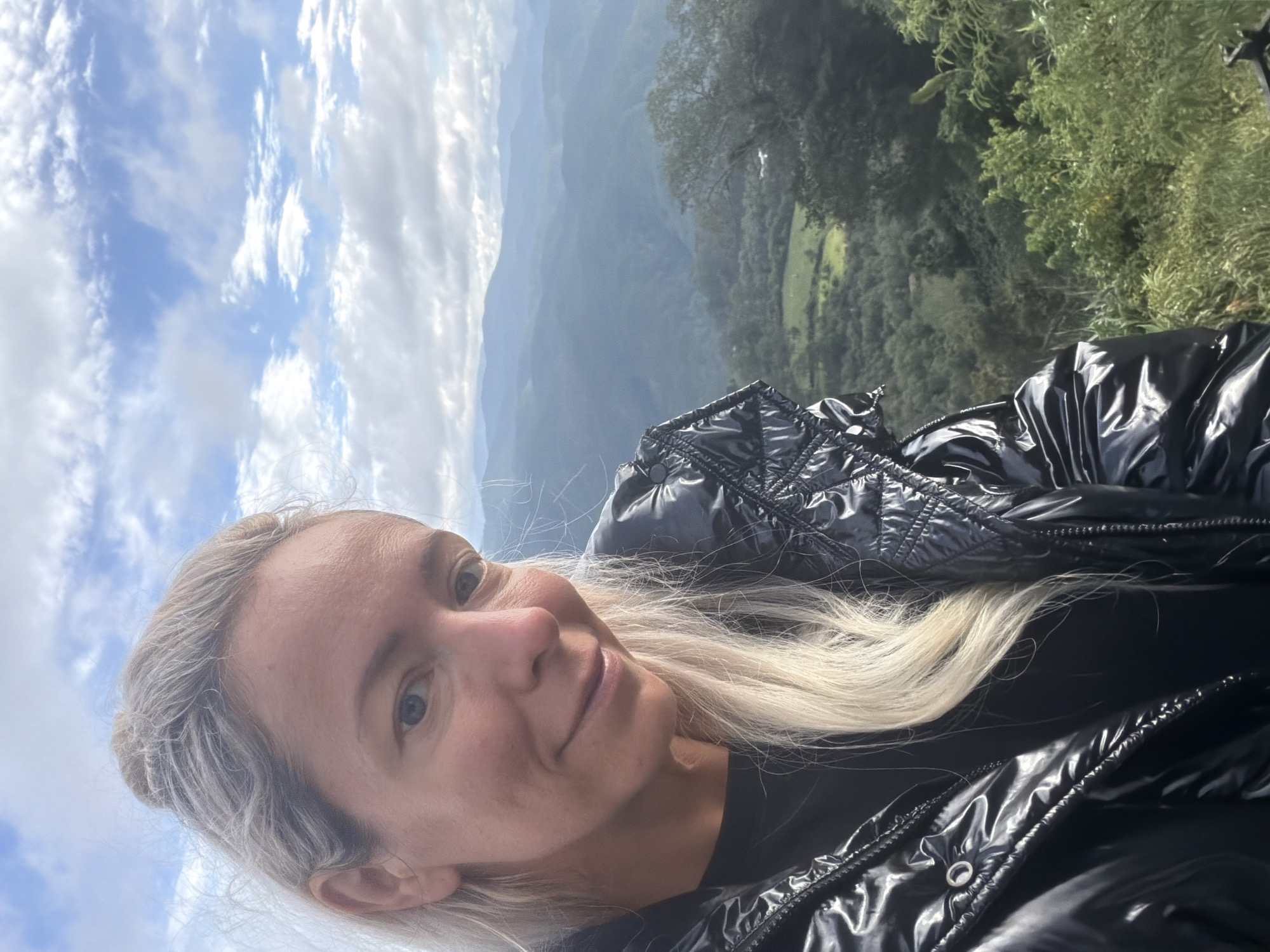 The image is a selfie taken outdoors. A blonde woman with blue eyes is in the foreground, looking at the camera and smiling. She is wearing a black puffer jacket and a black shirt. Behind her, there is a view of rolling hills covered in trees and vegetation, with a cloudy blue sky above. The woman's hair is light blonde and pulled back. The landscape suggests a mountainous or hilly region. The photo is taken from a high vantage point, looking down onto the valley. There is no information or visual cues in the image to suggest the location is in the city of Phát Chi, Vietnam.