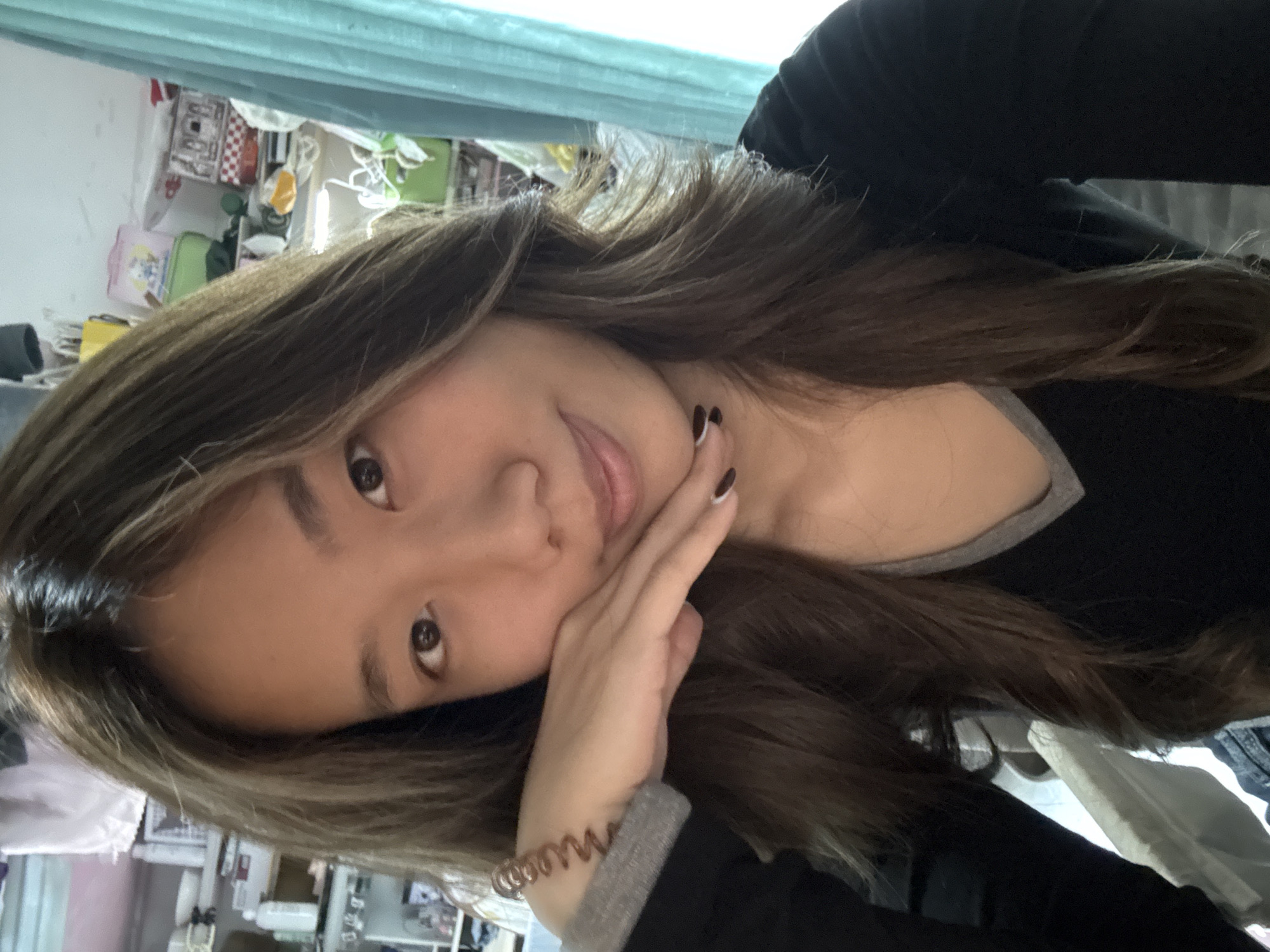 A young woman with long, highlighted brown hair is posing for a selfie. She is wearing a black top with a grey trim around the neckline and a spiral-shaped bracelet on her left wrist. Her nails are painted black with a white accent on one nail of her right hand. Her right hand is touching her face. The background appears to be a room with various items on a shelf or wall. The image is rotated, so the subject's head is tilted to the left. There is nothing to indicate this image was taken in the Nan'an District of China.