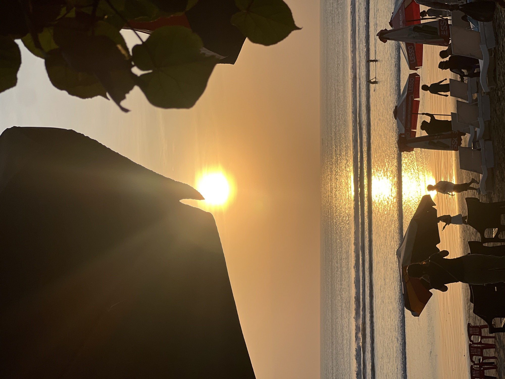 This is a vertical, medium-shot photograph of a sunset on Kuta Beach in Kuta, Indonesia. The sun is a bright, golden orb positioned slightly left of center in the sky, casting a strong reflection on the calm ocean waves. In the foreground, silhouettes of people are visible on the sandy beach, some standing under beach umbrellas and others near lounge chairs. Two beach umbrellas, one red and white and another with yellow and orange hues, are prominent. Several lounge chairs are arranged on the sand. The ocean has gentle waves rolling towards the shore, and a few individuals can be seen in the water, with one person appearing to be surfing or paddleboarding. The top left and right corners of the image are framed by dark, shadowy foliage and an abstract dark shape. The sky is a gradient of warm orange and yellow hues.