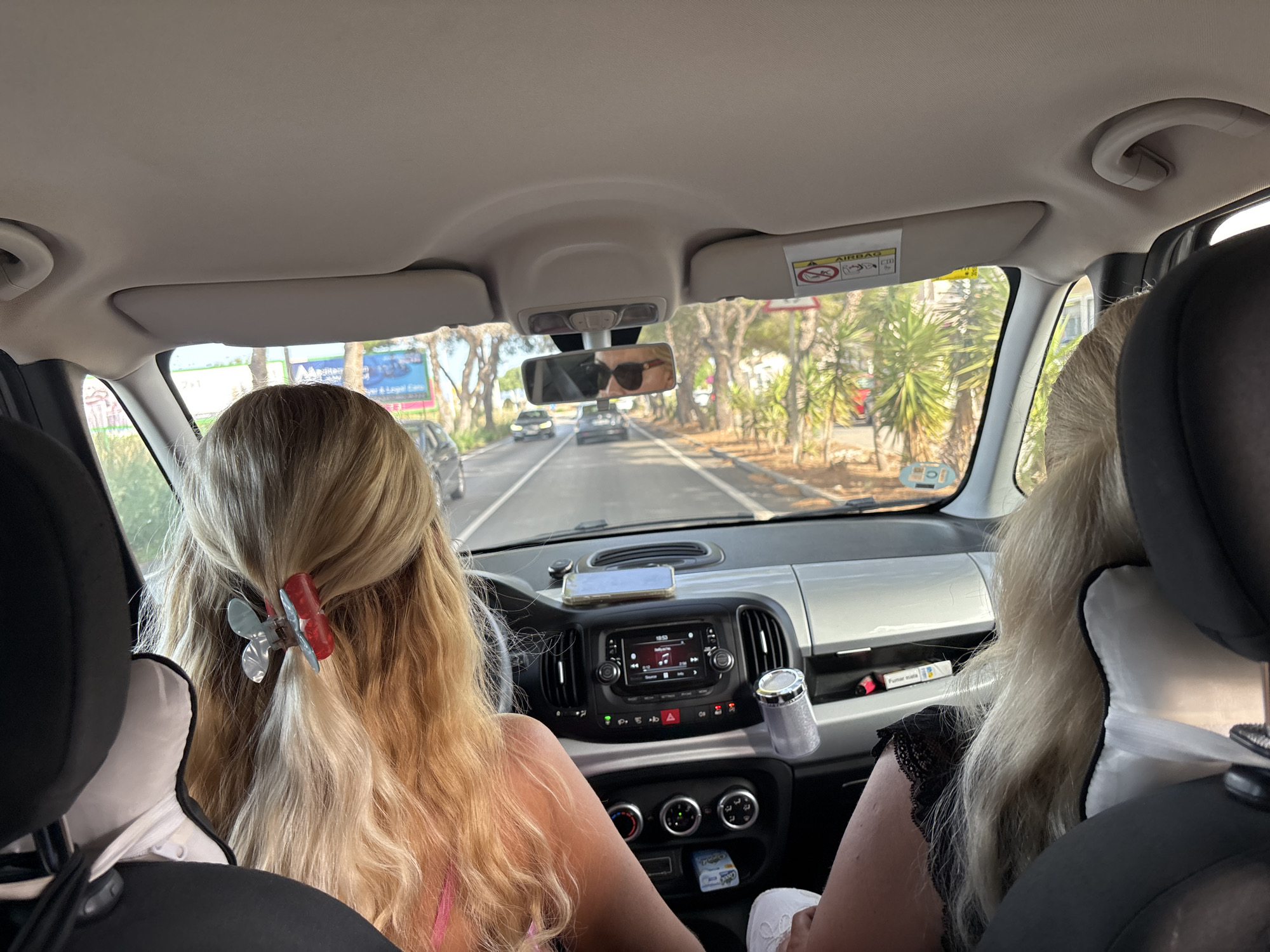 Two individuals, likely women based on their blonde hair, are inside a vehicle, viewed from the back seat looking forward. The driver, in the foreground on the left, has blonde hair styled with a blue and red butterfly hair clip. The person in the passenger seat on the right also has blonde hair. The car's interior is visible, including the dashboard with a radio display, air vents, and climate control knobs. A smartphone is mounted on the dashboard. Outside the car, a road lined with trees and palm trees is visible, suggesting a warm climate. There are other vehicles on the road ahead. A sun visor with an airbag warning sticker is also apparent. The scene appears to be a daytime drive. The question asks to include the location in the city of Orihuela, Spain, but this information cannot be determined from the image itself.