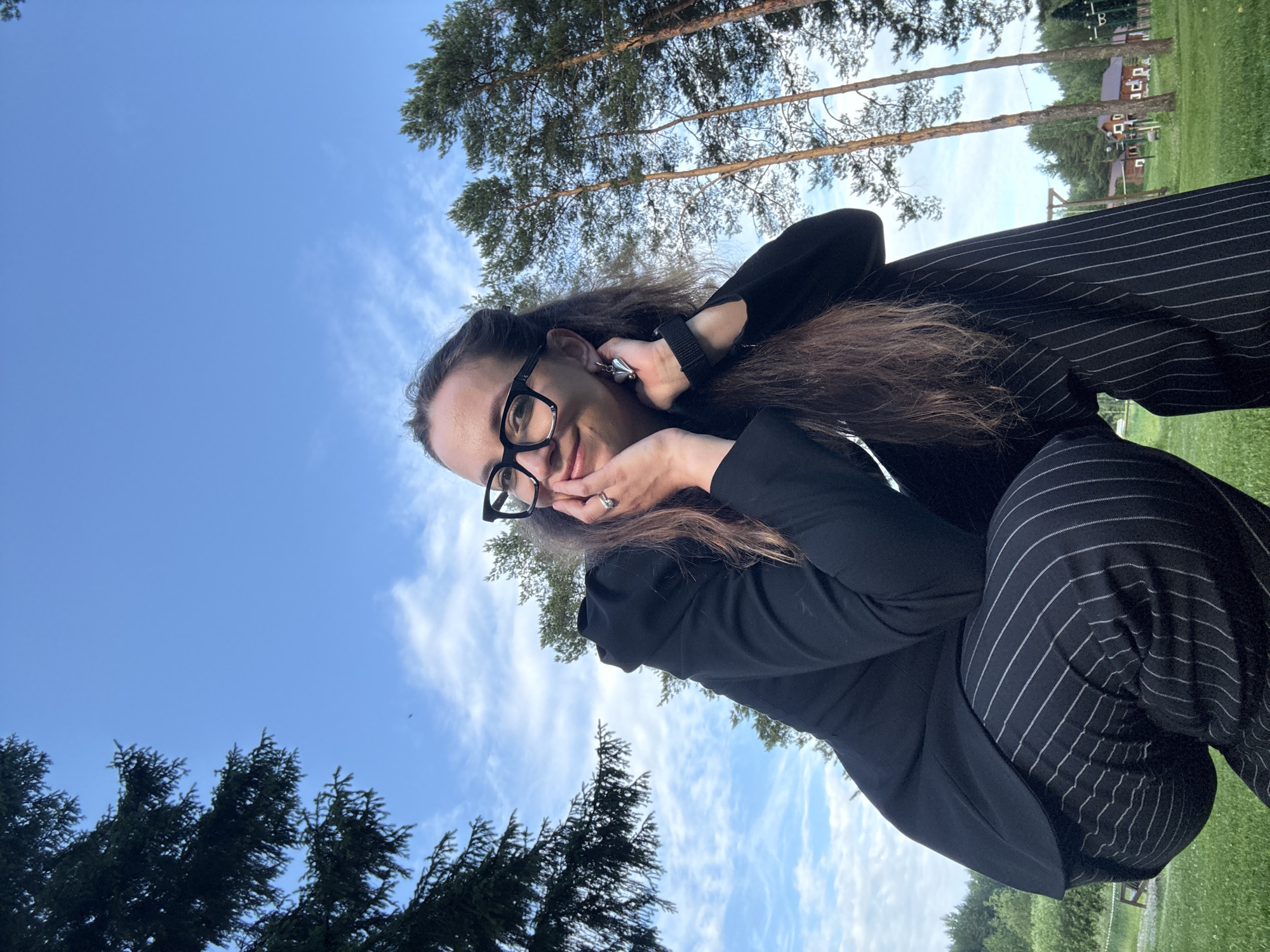 A medium shot captures a woman with dark, long, wavy hair and glasses, wearing a black blazer and pinstripe pants, crouching outdoors. Her hands are near her face, and she is looking upwards and to the left with a slight smile. The background features a bright blue sky with sparse clouds, tall pine trees, and a glimpse of houses and wooden structures in the distance. The setting appears to be a park or open grassy area.