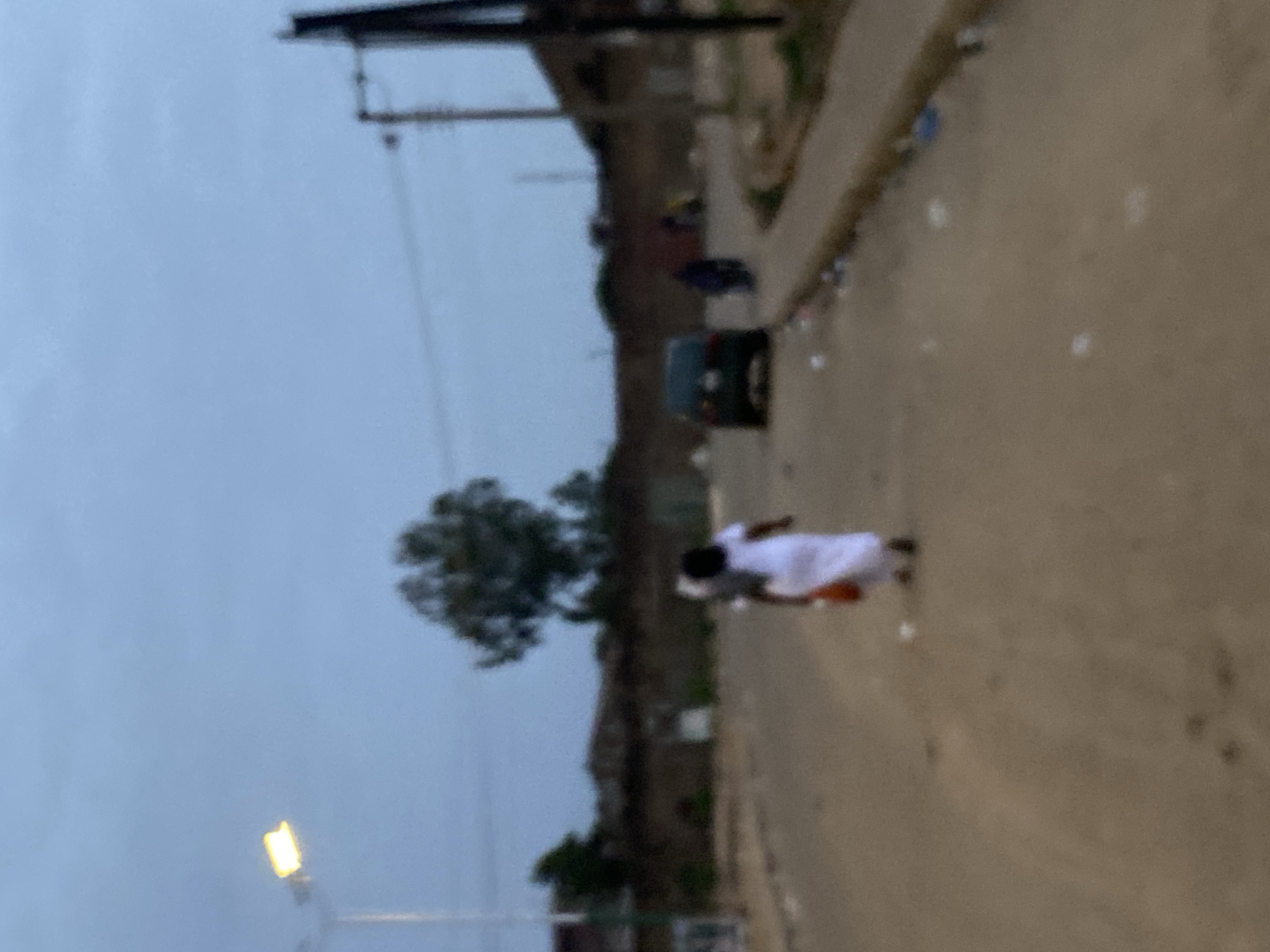 The image shows a dirt road in Bukuru, Nigeria. A person in white clothing is walking on the road. A black vehicle is parked on the side of the road. There is a tree and a utility pole visible in the background. The sky is overcast.
