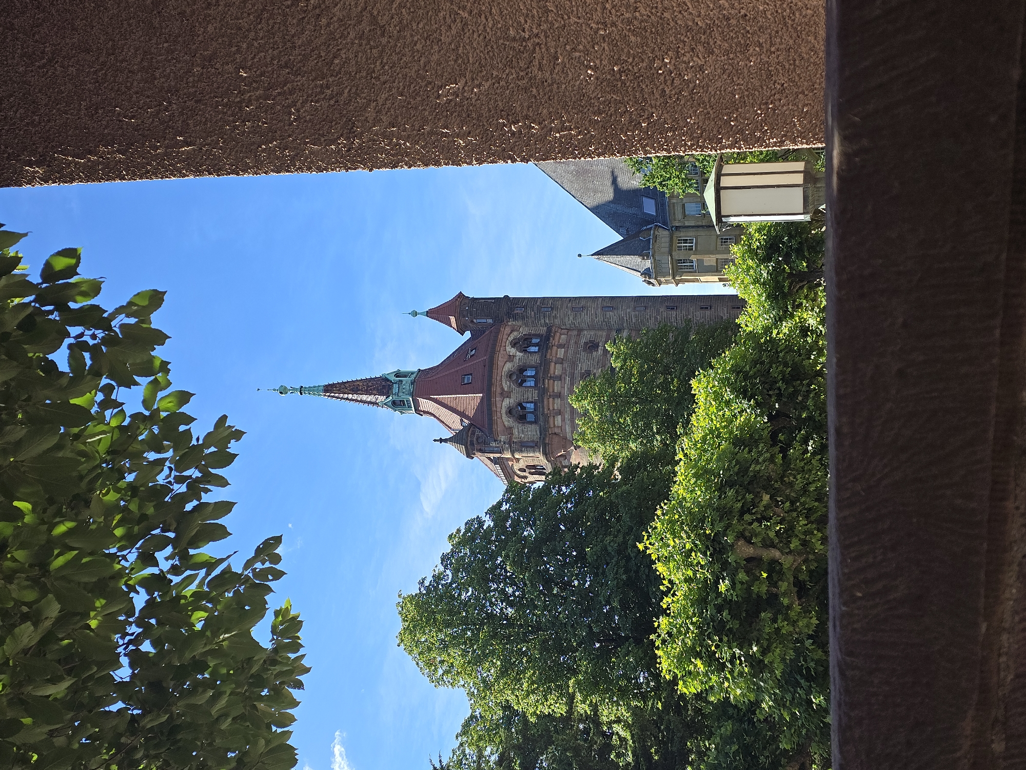 This is a photograph of the Worms Main Train Station building, a historic railway station in Worms, Germany, on a clear, sunny day. The image is framed by lush green foliage in the foreground, with leaves hanging from the top and sides. A textured, earthy brown wall or structure occupies the right side of the frame.
Dominating the center and left of the image is the distinctive architecture of the train station. A tall, multi-story tower with a conical, reddish-brown roof and copper-colored accents stands prominently. It features arched windows and a dark stone facade. Adjacent to this main tower, a smaller, similarly styled tower with a pointed roof can be seen. In the background, another building with multiple stories and a mansard roof is visible.
A modern-looking, white outdoor lamp with a copper-colored top is positioned in front of the station buildings, to the right of the main tower and behind some trees. The sky is a vibrant, cloudless blue, indicative of good weather. The overall scene captures a blend of natural greenery and historical architecture under a bright sky.