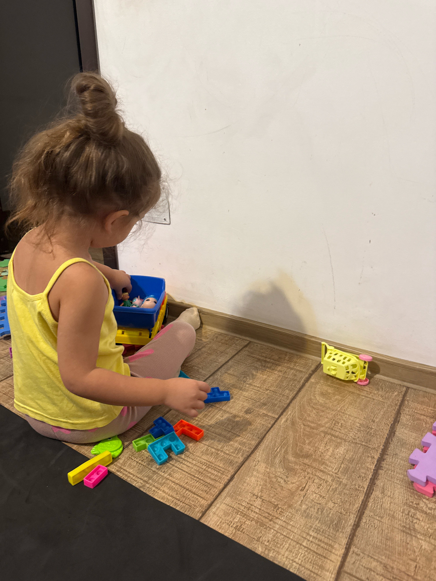 A young girl with her hair in a bun, wearing a yellow tank top and striped leggings, sits on a wooden floor playing with toys. She is reaching into a blue toy truck filled with small figures. Scattered around her are colorful building blocks, including a yellow and pink set and a green frog-shaped toy. To her right, a yellow toy shopping cart with pink wheels lies on its side near a white wall. In the background, a portion of a dark doorway is visible. The scene appears to be an indoor domestic setting.
