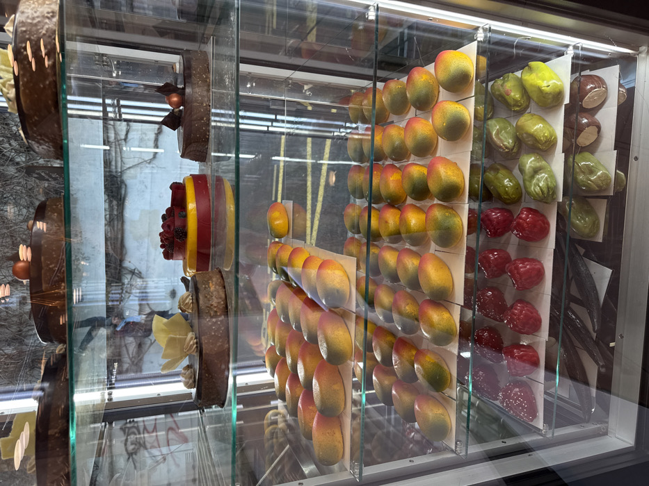 A display case features an array of desserts. Round chocolate cakes with varying decorations are positioned on the left. Multiple rows of fruit-shaped treats, including mangoes, green fruits, and raspberries, are neatly arranged on stands. Vanilla beans are visible to the right. The glass case reflects an outdoor scene, partially obscuring the desserts.