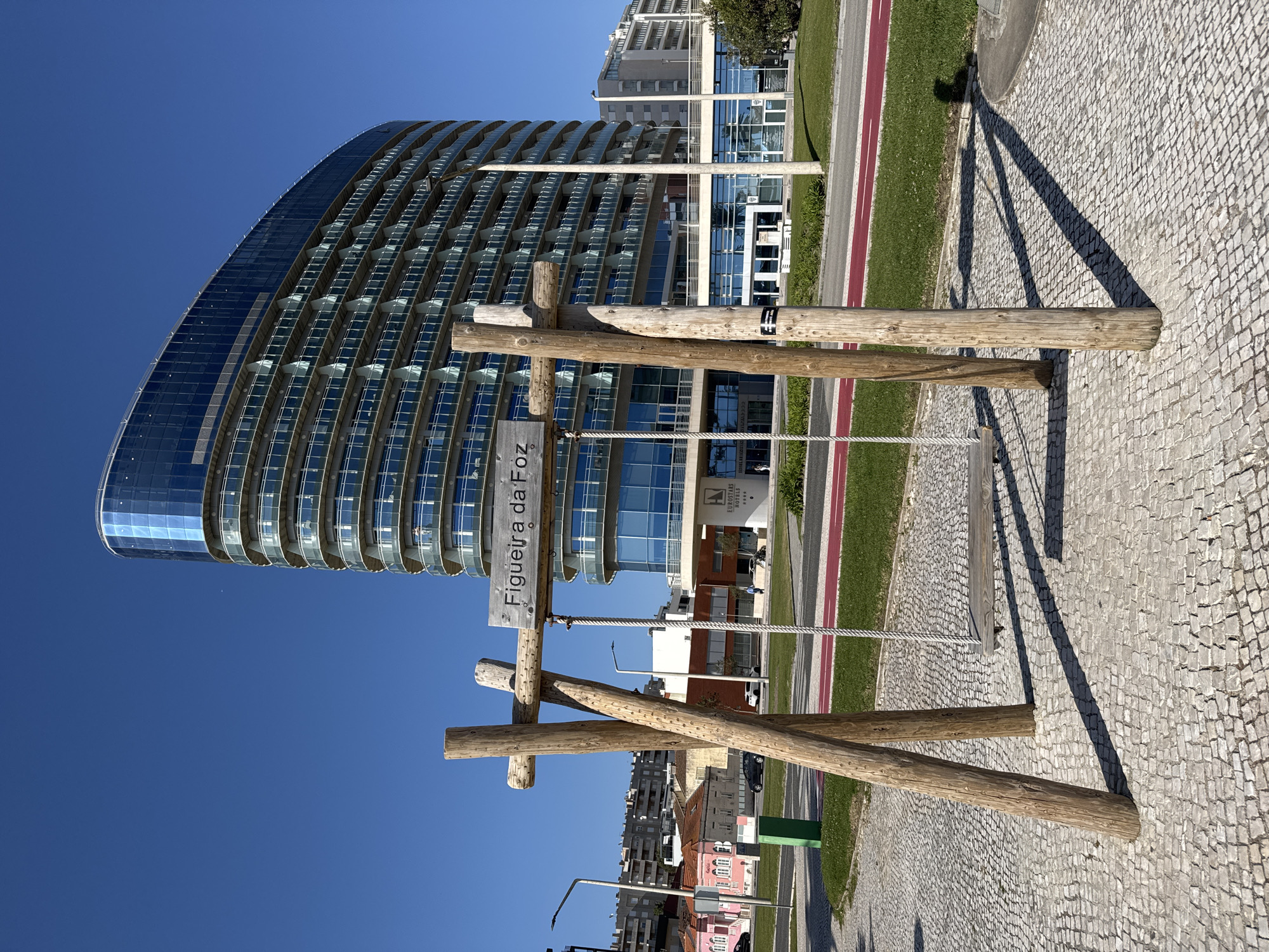 The image captures a sunny day in Figueira da Foz, Portugal. A wooden structure with a sign that reads "Figueira da Foz" stands prominently in the foreground. Behind it is a modern, curved glass building with many floors, possibly a hotel or apartment complex. To the right of the wooden structure, there is a red cycling path, followed by a patch of green grass. The ground is covered with cobblestones. The sky is clear and blue. Some buildings are visible in the far background.