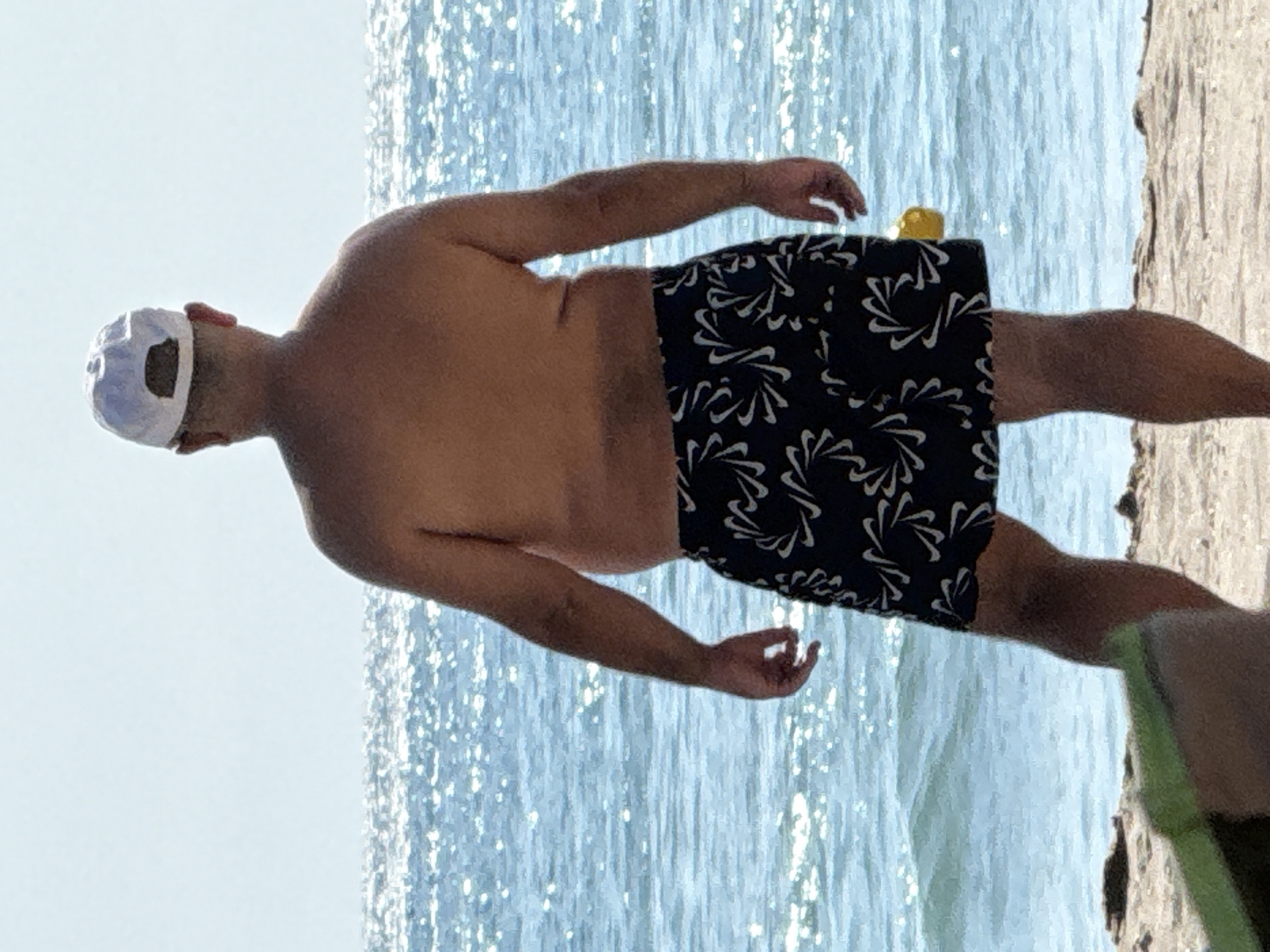 A man wearing a white baseball cap and dark swim trunks with a white pattern stands on a beach, facing away from the camera. He is shirtless, exposing his back and arms. The ocean is visible behind him, with shimmering sunlight reflecting off the water. The background is a clear, light blue sky. The man appears to be an adult.