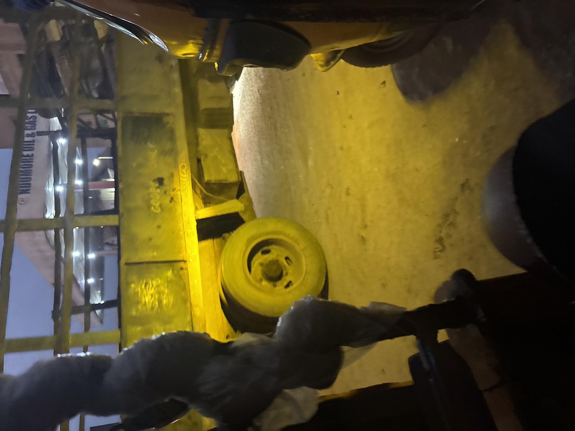 The image depicts a yellow truck with the signage "NDUMORE OIL & GAS" visible at the top. The truck is parked on a street at night, illuminated by yellow streetlights, casting a warm glow on the surroundings. In the foreground, a person's legs are visible, wrapped in what appears to be grey fabric. To the right, the front of a yellow vehicle, possibly a tuk-tuk or a smaller car, is also visible. The scene is likely located in Onitsha, Nigeria, a bustling commercial city known for its vibrant street life and vehicular traffic. The overall impression is one of a typical night scene in a Nigerian city, with the presence of commercial vehicles and people engaged in their daily activities.