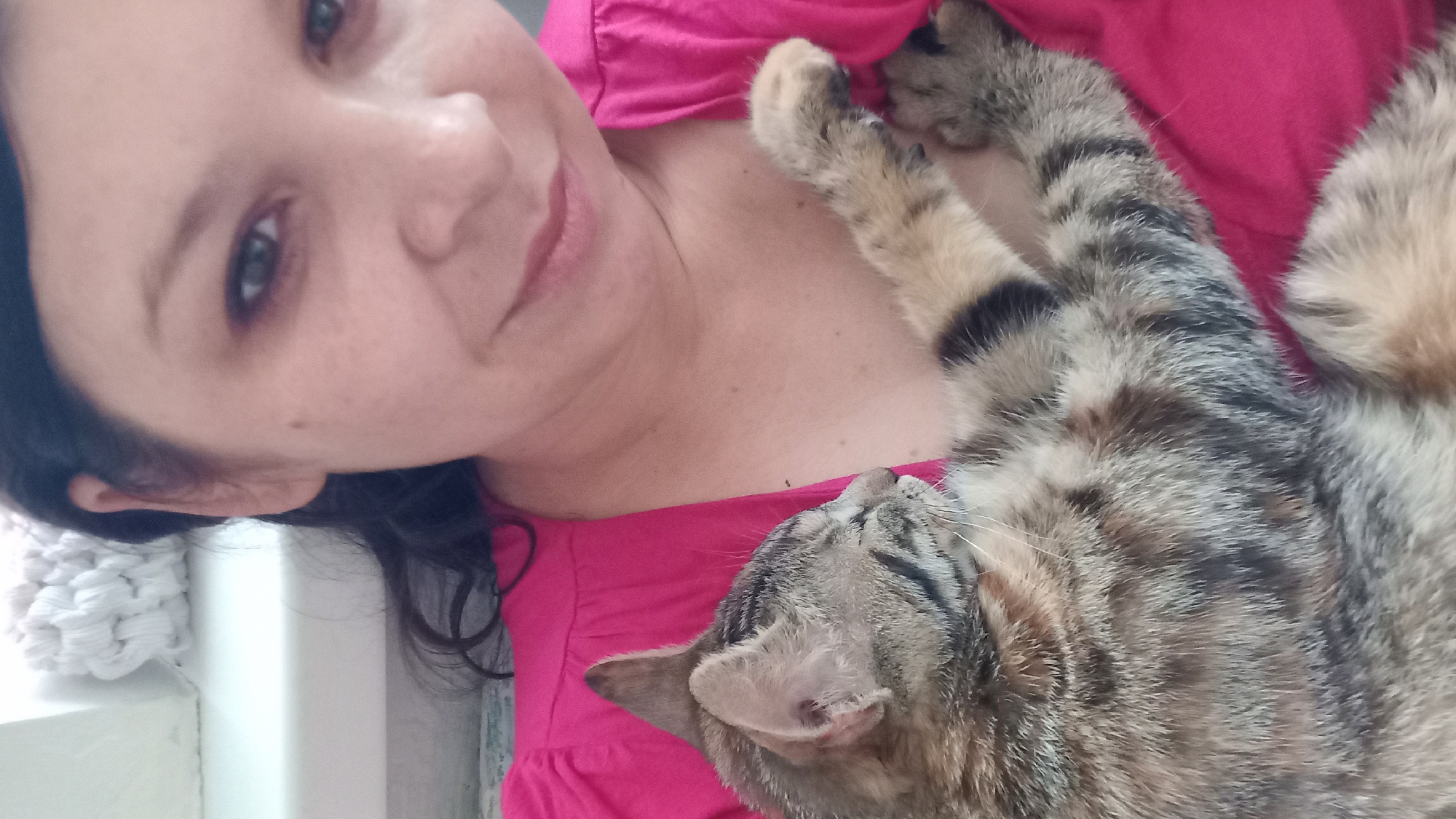 A woman is holding a tabby kitten in her arms. The woman is wearing a bright pink shirt. The kitten is sleeping and resting its head on the woman's chest. The background is out of focus, but it appears to be a home environment. The lighting is natural, coming from the left side of the image.