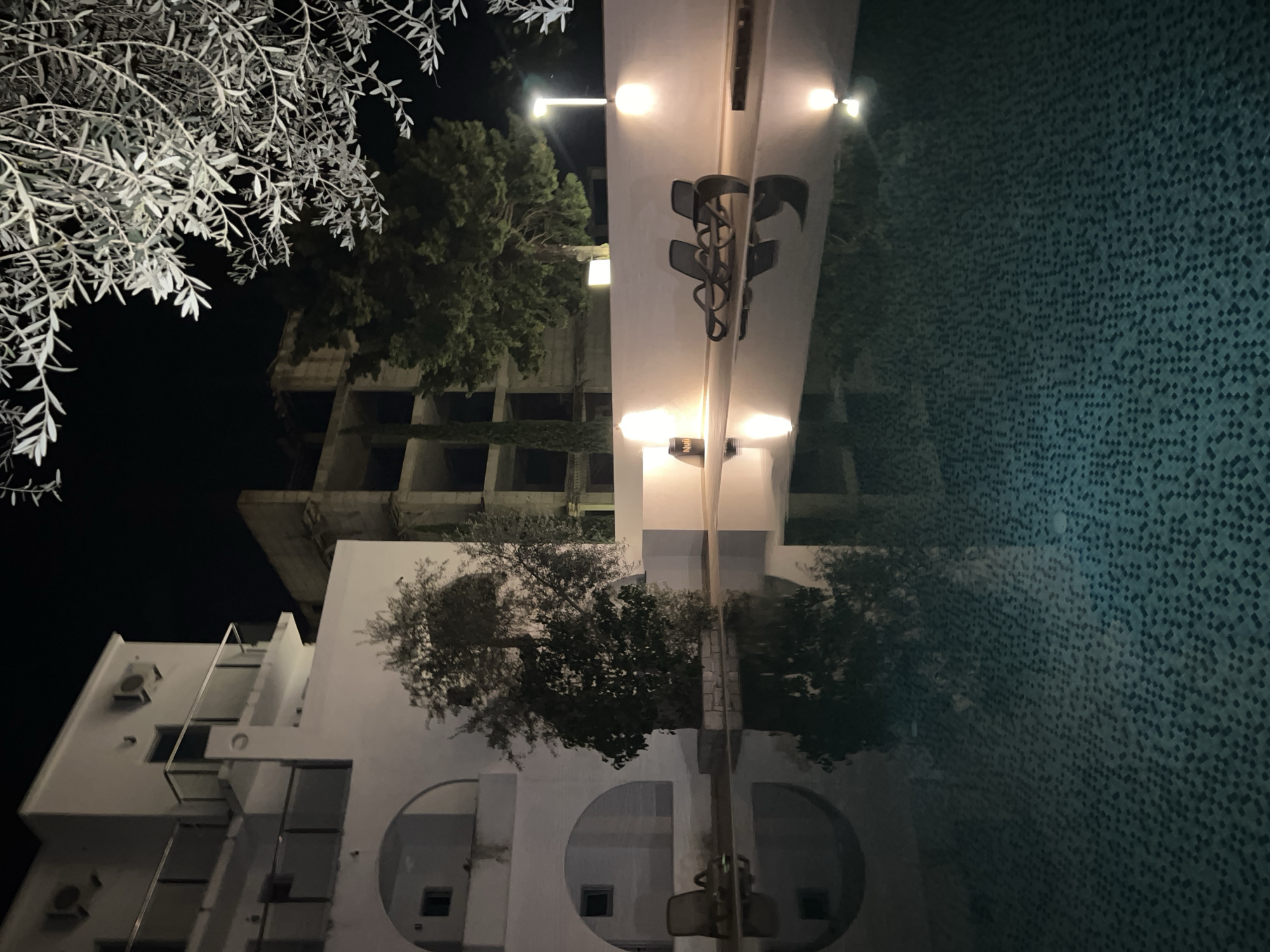 The image shows a night view in Brežine, Montenegro. A multi-story building with white walls, balconies, and air conditioning units is on the lower left. Above it and slightly to the right is a taller, unfinished concrete building with multiple window openings. A tree with sparse, light-colored foliage is in the upper left corner of the frame. In the center, a brightly lit structure reflects in the water of what seems to be a pool. A tree is also reflected in the pool’s surface. The pool's tiles are visible. Overhead lights illuminate the central structure. A partially visible sign is present on the lit structure.