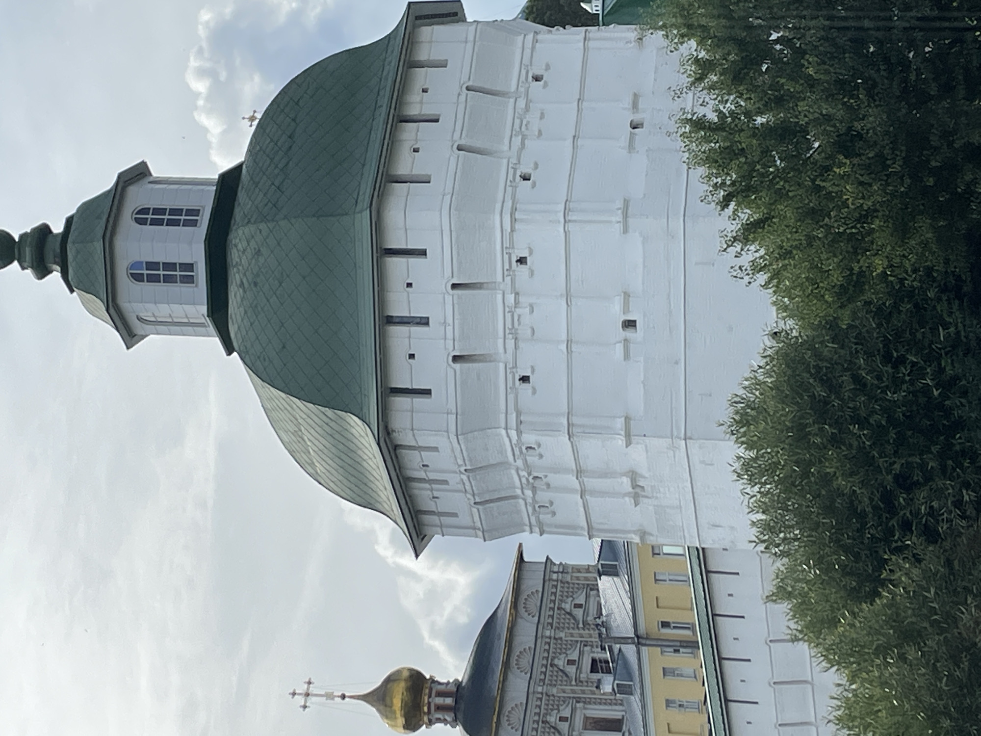 This is a close-up, eye-level, outdoor, daytime photograph of the Trinity Lavra of St. Sergius in Sergiyev Posad, Russia. The main subject is a large, white, multi-tiered circular tower with a green, segmented domed roof. The tower has numerous small rectangular openings and decorative horizontal banding. To the left of this tower, a portion of a building with a golden dome topped by a cross is visible. This building features intricate decorative patterns on its facade and a yellow lower section. Lush green trees are in the foreground, partially obscuring the base of the white tower and the lower walls of the complex. The sky is overcast with scattered clouds. There are no people visible in the frame.