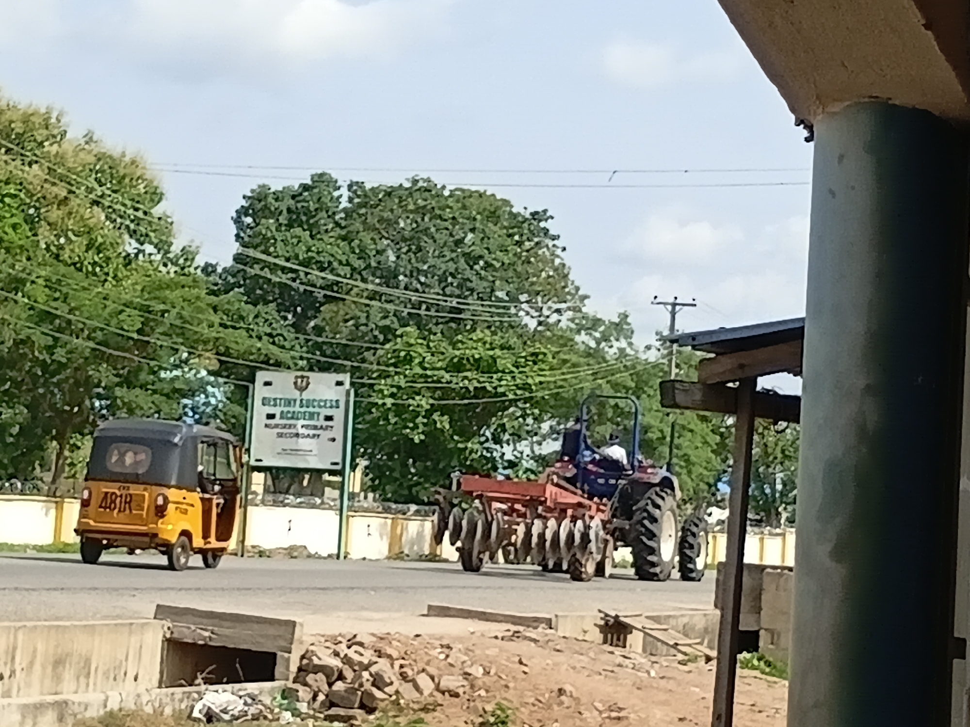 In the city of Jalingo, Nigeria, on a paved road, a yellow and black auto-rickshaw with the license plate "481R" is parked to the left. In the background, a white sign for "Destiny Success Academy" with "NURSERY, PRIMARY, SECONDARY" is visible. To the right of the auto-rickshaw, a blue tractor with a red plow attachment is driving on the road. A person is seated in the driver's seat of the tractor. The scene is framed by a concrete structure in the foreground on the right and trees and a blue sky with scattered clouds in the background.
