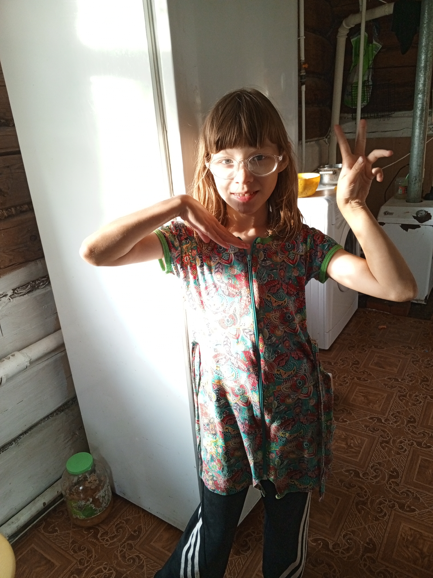 A young girl with long brown hair and glasses stands in front of a large white appliance, possibly a refrigerator or washing machine. She is wearing a colorful, patterned short-sleeved dress with a zipper down the front and dark pants with white stripes. She is making a peace sign with her right hand, and her left hand is raised near her face. The room appears to be a utility area, with pipes, a washing machine, and a large glass jar with a green lid on the floor to the left. The floor is tiled with a brown, patterned design. Sunlight streams in from the left, illuminating the scene and casting shadows. The overall impression is one of a casual, indoor snapshot taken during the day.