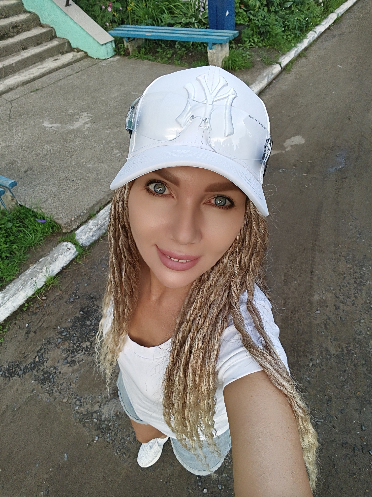 An eye-level, full-body selfie taken from a slightly high angle shows a blonde woman with braided hair wearing a white t-shirt, light-colored shorts, and a white New York Yankees baseball cap. She is outdoors in what appears to be a park or public space, with a concrete walkway, patches of grass, and a blue bench visible in the background. The woman is smiling and looking directly at the camera.