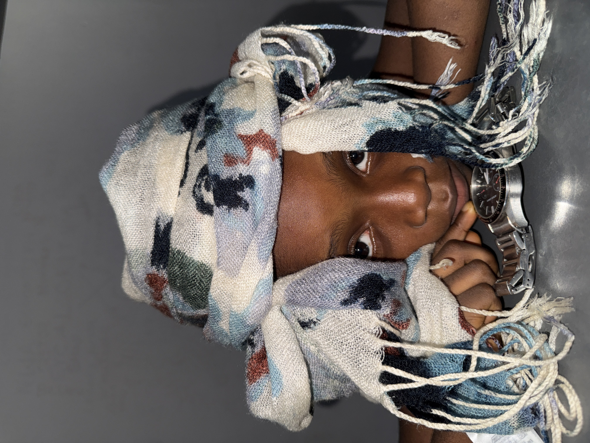 A close-up portrait shows a person of color, likely female, positioned against a gray background. A patterned, loosely woven scarf is wrapped around the person's head and upper body, partially obscuring their face. Only one eye and part of the nose and mouth are visible. A silver wristwatch is visible on the person's wrist, which rests against their cheek. There is no information regarding the location of Ado, Nigeria.