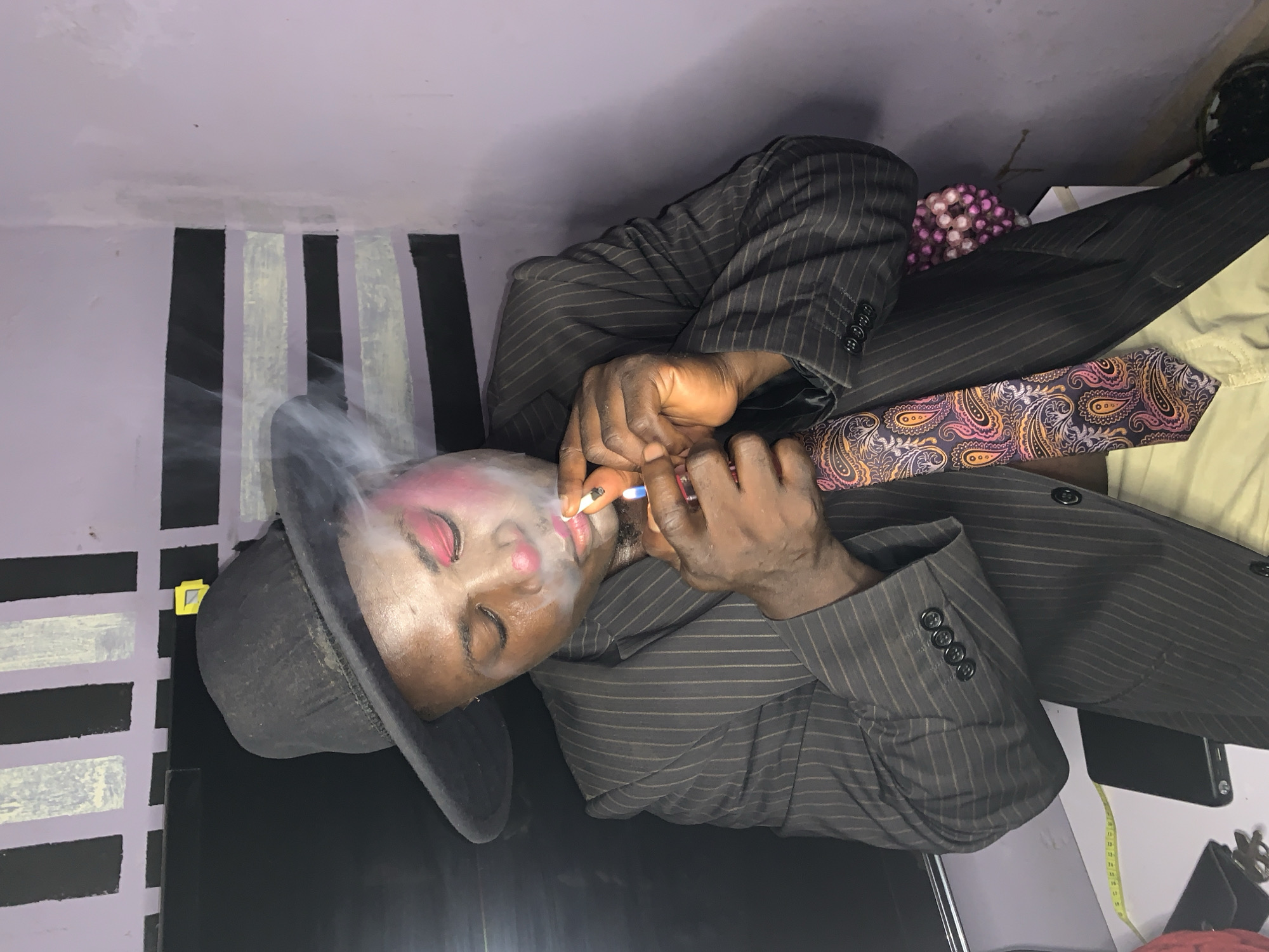 In Akwanga, Nigeria, a person with clown-like makeup is seen wearing a black hat and a pinstripe suit. They are holding a lit cigarette in their right hand and appear to be inhaling from it, with smoke rising from the cigarette and around their face. The person's eyes are closed, and their face has pink blush on the cheeks and a red nose. A colorful paisley tie is worn with the suit. The background features a wall with a pattern of black and white stripes. The scene is illuminated by artificial light.