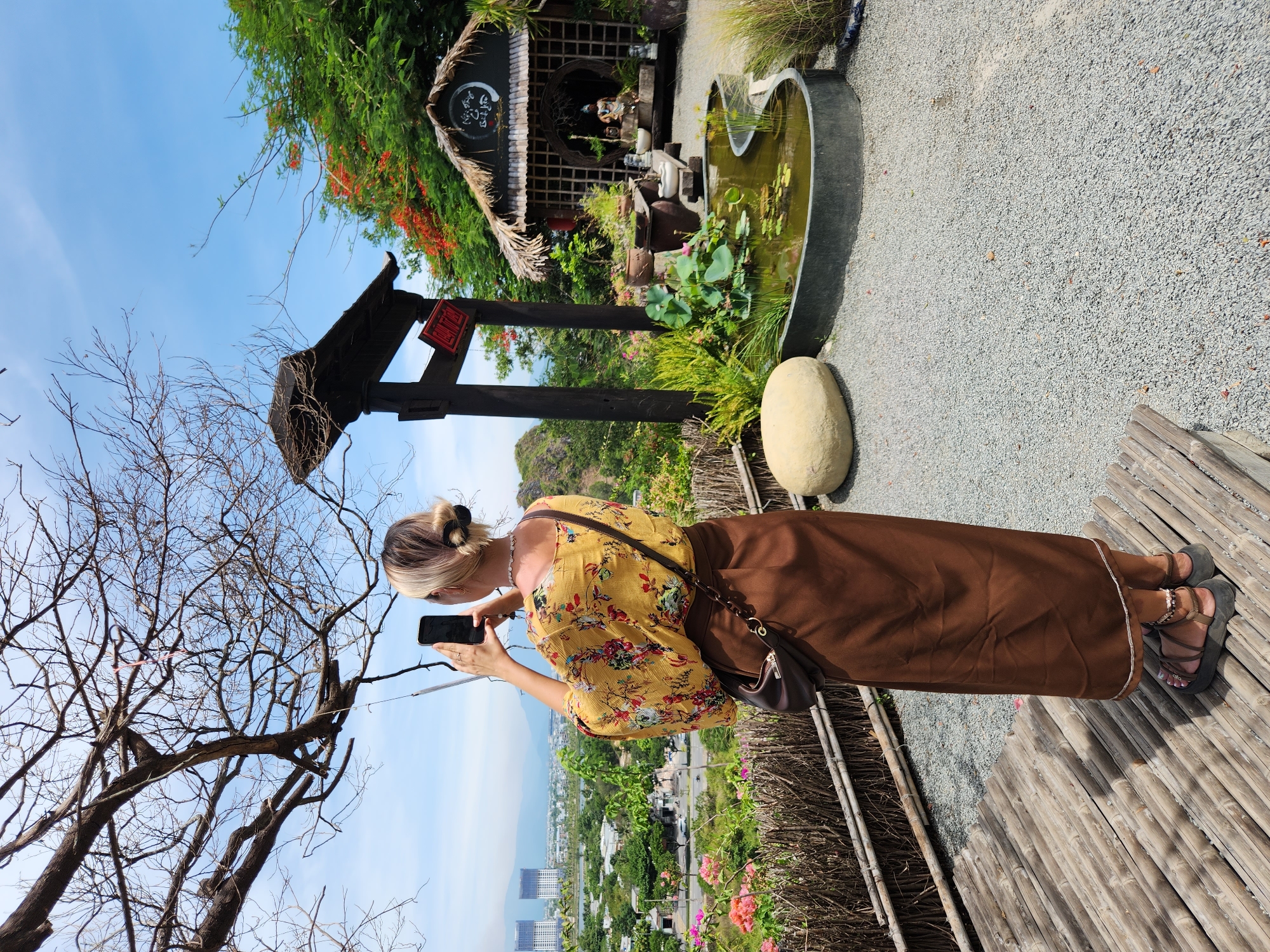 A woman with blonde hair pulled up in a bun is standing on a bamboo platform, holding a smartphone in her right hand and taking a picture of a Japanese-style garden. She is wearing a yellow top with a floral pattern and a long brown skirt. A dark brown purse is slung across her body.
The garden features a small pond with lily pads, surrounded by lush greenery and potted plants. A wooden structure with a thatched roof is visible in the background, housing a statue and other decorative items. A dark wooden torii gate stands to the left of the garden, with a red sign attached to it. In the distance, the city skyline of Đà Nẵng, Vietnam, is visible, with tall buildings and mountains. The sky is clear and blue. The ground is covered with gray gravel.