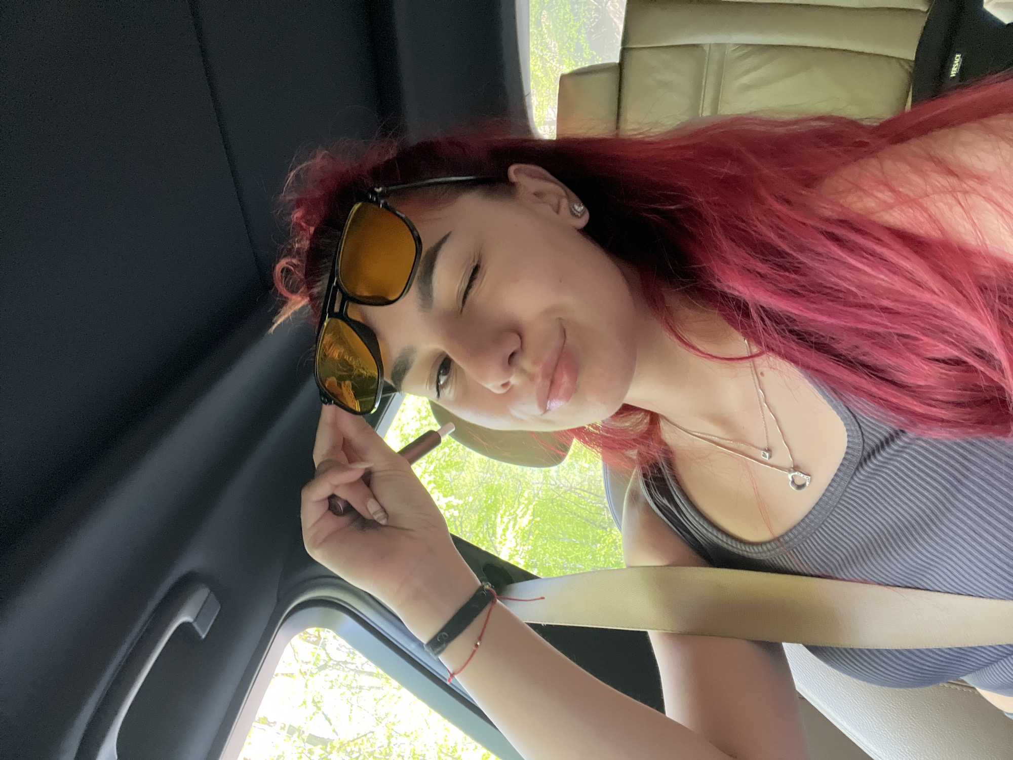 A young woman with long, vibrant red hair is featured in a close-up selfie taken from inside a car, presumably in Vrata, Bulgaria. She is positioned diagonally in the frame, winking with her right eye and displaying a playful, slight smile. She wears a grey ribbed tank top and a layered silver necklace featuring a small square pendant and an open heart-shaped pendant. Large yellow-tinted sunglasses rest on her head, and she is holding one arm of the sunglasses with her left hand. Her left wrist is adorned with a black and a red string bracelet.
The setting is the interior of a car, characterized by a dark overhead panel, a grey grab handle, and light-colored leather seating. A beige seatbelt diagonally crosses her chest. Through the car window, bright green foliage is visible, suggesting a sunny, daytime environment. A black item, possibly a bag or garment, is seen in the background on the seat, with the brand name "VERSACE" clearly visible.