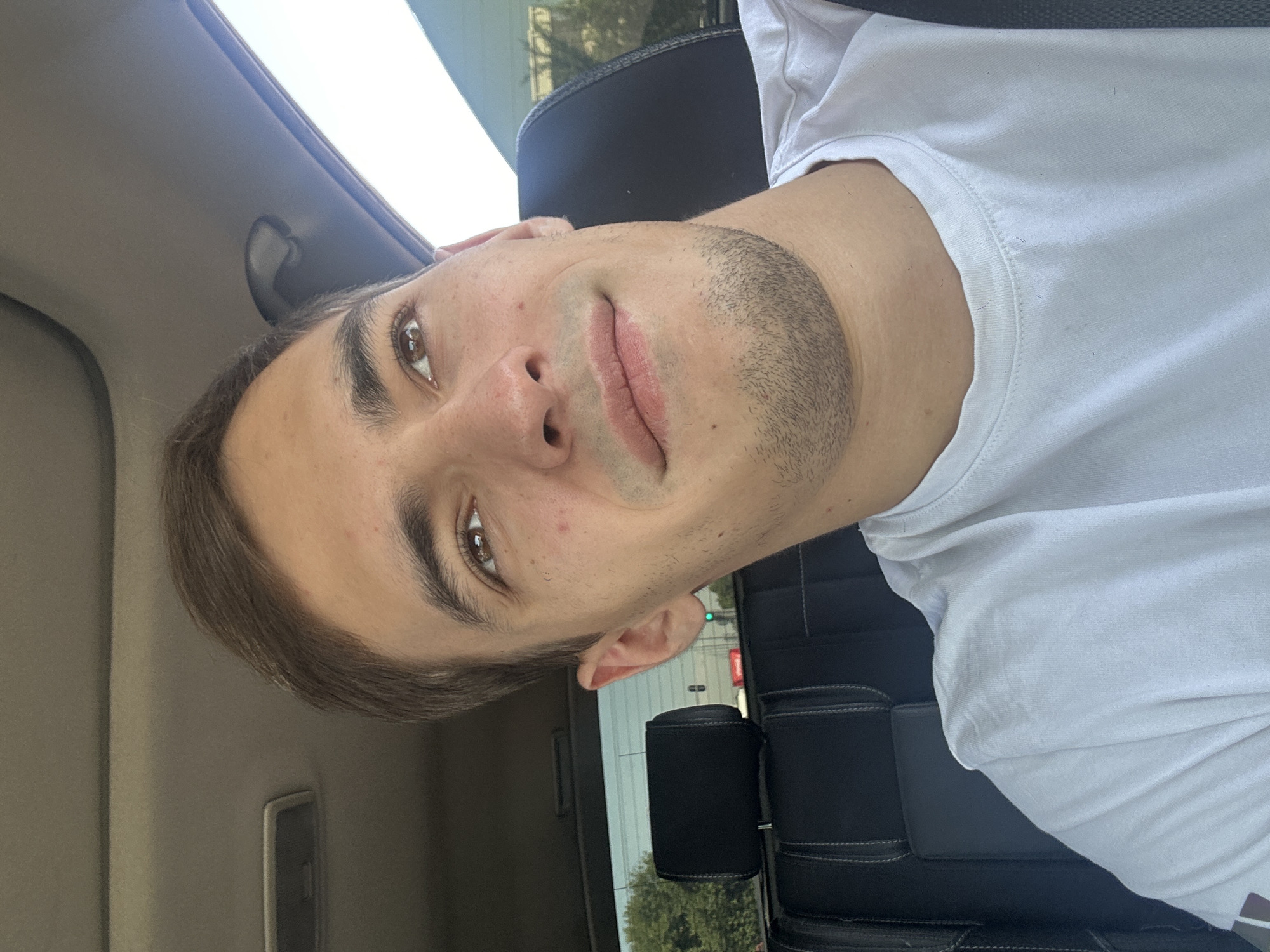 A young man with light brown hair and a light beard is sitting in the passenger seat of a car, looking up and to the right with a slight smile. He is wearing a white t-shirt. The interior of the car is visible, including a black seat with white stitching and a part of the car's roof liner. Outside the car, trees and traffic lights are visible through the rear window, suggesting an urban environment. The lighting indicates it is daytime. This scene is not identifiable as being in Tashkent, Uzbekistan.