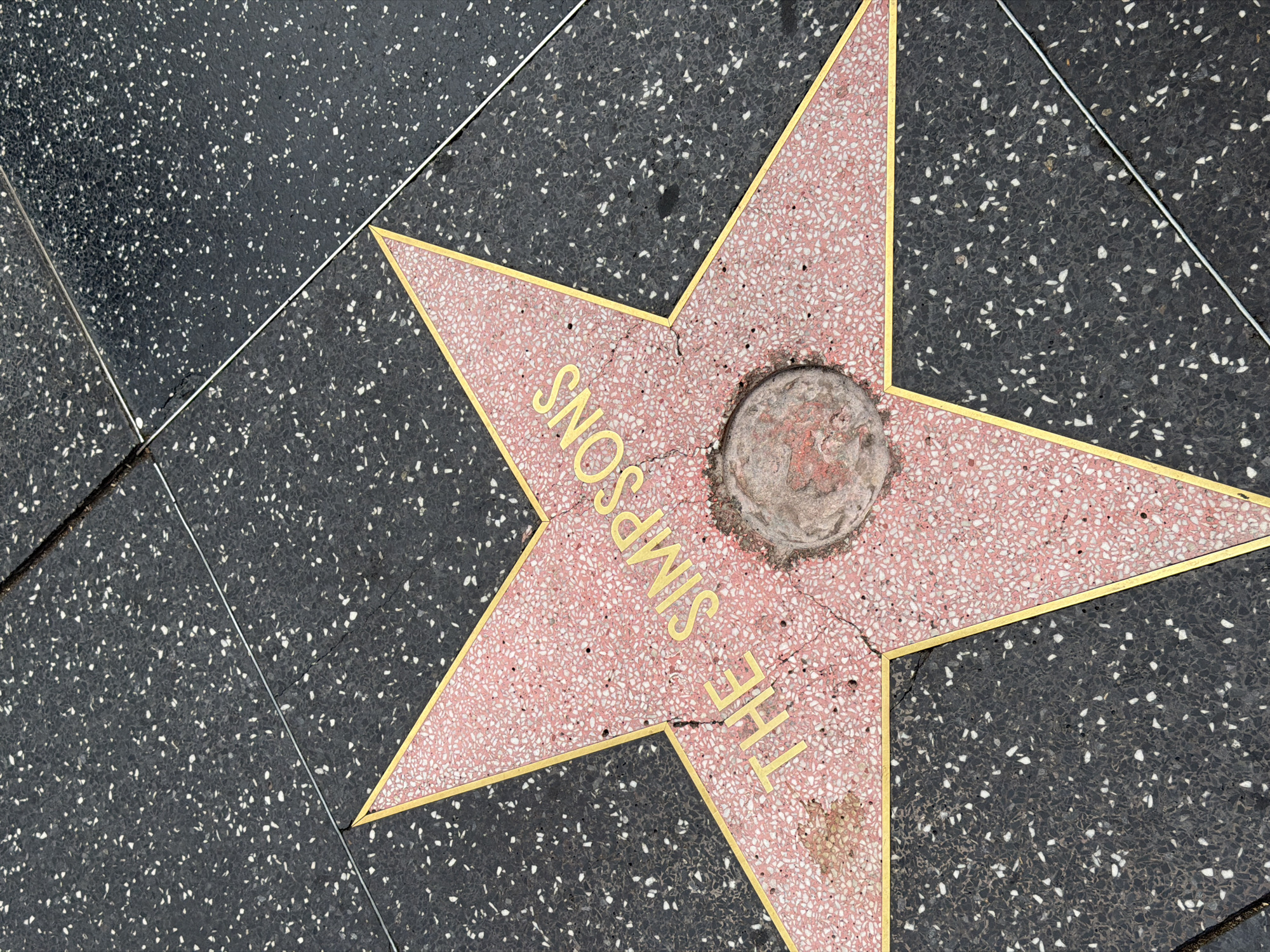 The image shows the Hollywood Walk of Fame star for "The Simpsons," located in Los Angeles, California. The star is pink with gold lettering and trim, embedded in a dark gray terrazzo sidewalk. The central medallion is worn and discolored. Cracks are visible in the star.