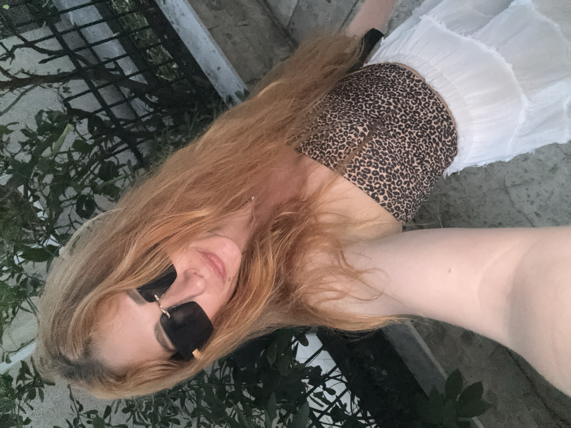 A medium-shot, high-angle selfie shows a young woman with long, reddish-blonde hair and fair skin. She is wearing large, black, rectangular sunglasses and a leopard-print strapless top, paired with a white, ruffled skirt. She is outdoors, with green foliage and a black metal fence visible in the background, suggesting a residential or garden setting. The lighting suggests it is either early morning or late evening.
