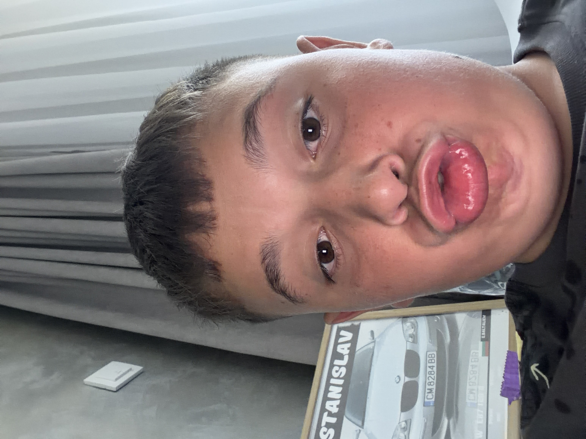 The image is a close-up, selfie-style photograph of a young person with short, dark hair and a fair complexion. The person is making a duck face, with their lips pursed. They have brown eyes and light eyebrows. Freckles are visible on their cheeks.
In the background, to the left, there is a framed picture of a silver BMW car with a Bulgarian license plate that reads "CM 8284 BB". The name "STANISLAV" is visible at the top of the frame. The picture is propped up against a grey wall with a white light switch.
To the right of the person, a grey curtain is visible, partially obscuring a white wall or window. The overall setting appears to be indoors, possibly a bedroom or living area. There is no information in the image to suggest a specific location within the city of Zmeitsa, Bulgaria.