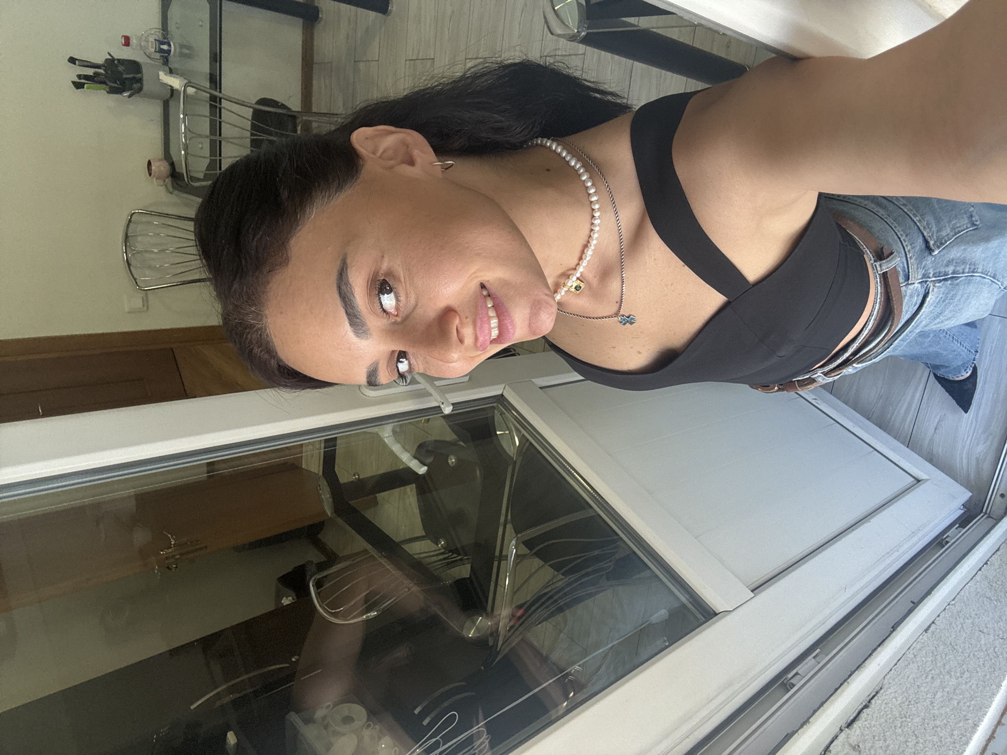 A woman with dark hair and brown eyes is taking a selfie in what appears to be a domestic interior, possibly an apartment in Tbilisi, Georgia. She is wearing a black crop top, jeans, and a pearl necklace with two pendants. The background shows a glass door reflecting a room with a table, chairs, and some items on the table. The floor is tiled. The woman is smiling and looking at the camera.