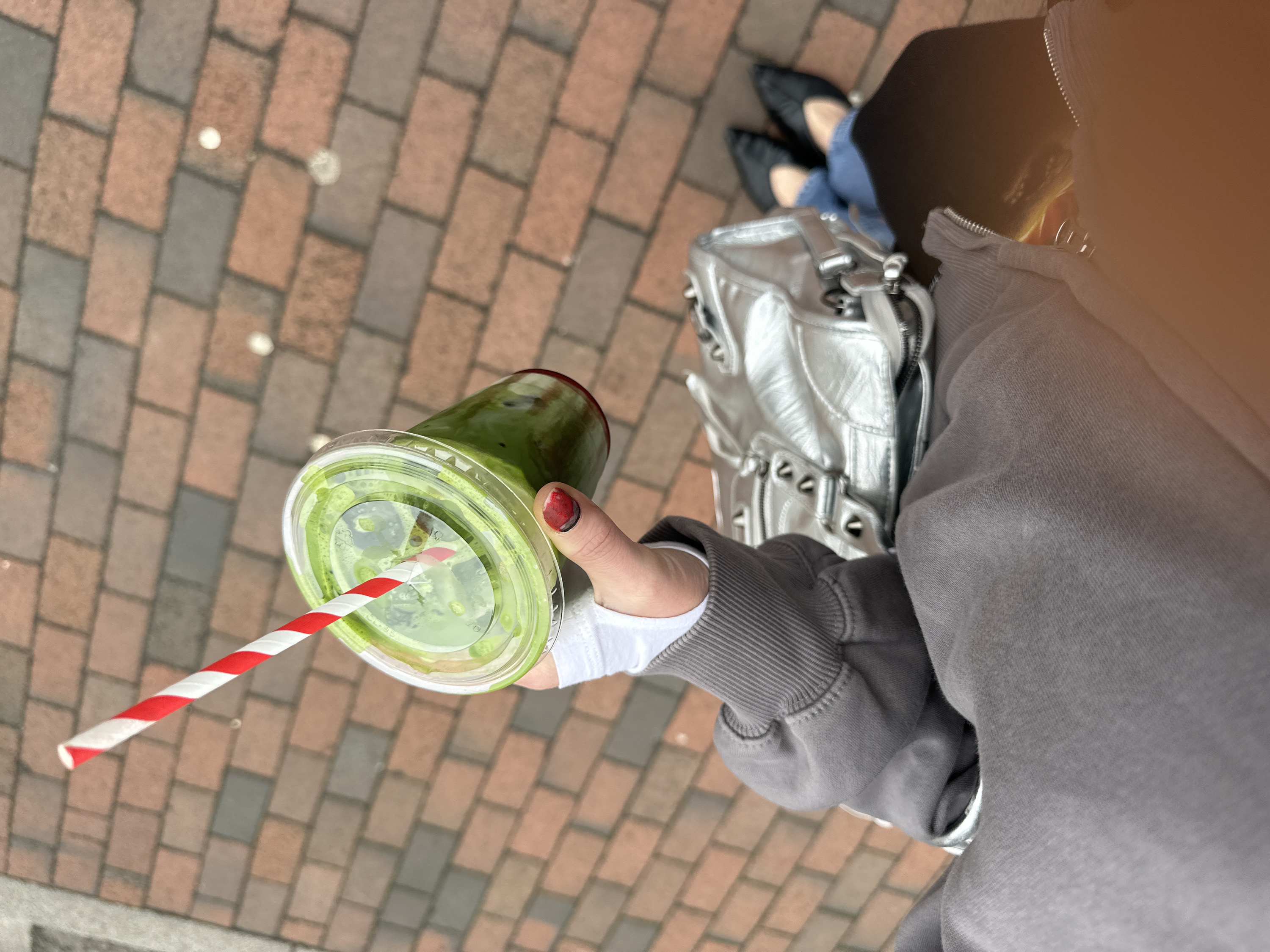 An overhead, close-up view depicts a person from the lower torso downwards, standing on a brick-paved surface in Tunbridge Wells, United Kingdom. The individual wears a grey hooded sweatshirt with ribbed cuffs, blue jeans, and black pointed-toe flat shoes. A silver metallic handbag, detailed with studs and zippers, is visible on their left side. The person's right hand, featuring a dark red and lighter-tipped painted thumbnail, holds a clear plastic cup containing a layered green beverage. A red and white striped paper straw is inserted into the drink. The ground is composed of a patterned arrangement of reddish-brown and darker grey rectangular paving bricks.