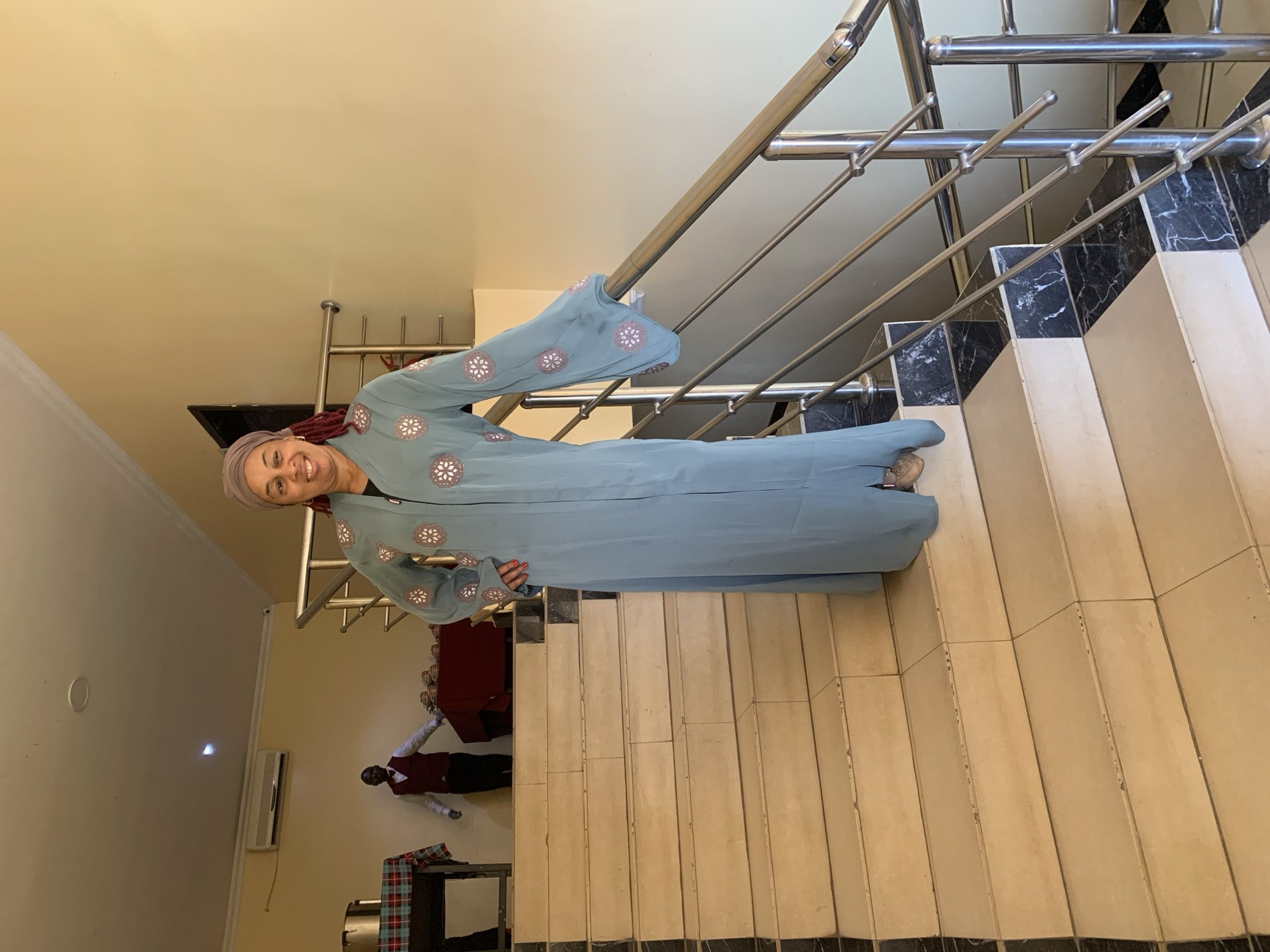 A woman stands on a tiled staircase with a metal railing inside a building. She is wearing a light blue, ankle-length garment with floral embroidery and a matching head covering. A man in a maroon uniform is visible standing near furniture in the background. The location is Gura Top, Nigeria.